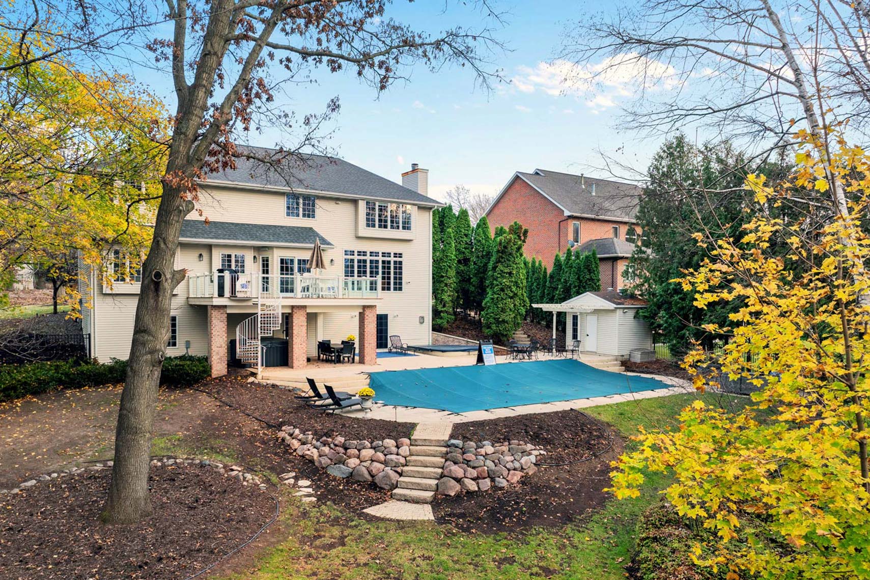 Residential real estate photograph featuring a two-story home with expansive windows, a covered swimming pool, patio furniture, and autumn foliage.