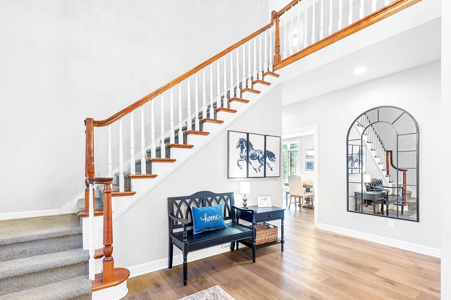 Real estate photograph of a spacious residential foyer featuring a wooden staircase and black bench, adorned by an arch mirror reflecting the interior. Blue bench cushion, hardwood flooring, gray carpeted stairs, and natural light streaming from an adjoining room enhance the inviting atmosphere.