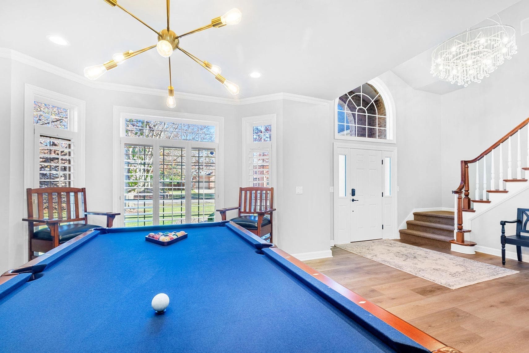 Residential interior space featuring a blue pool table under a modern overhead light fixture, with wooden chairs arranged by expansive windows and a staircase adjacent to the double-door entry.