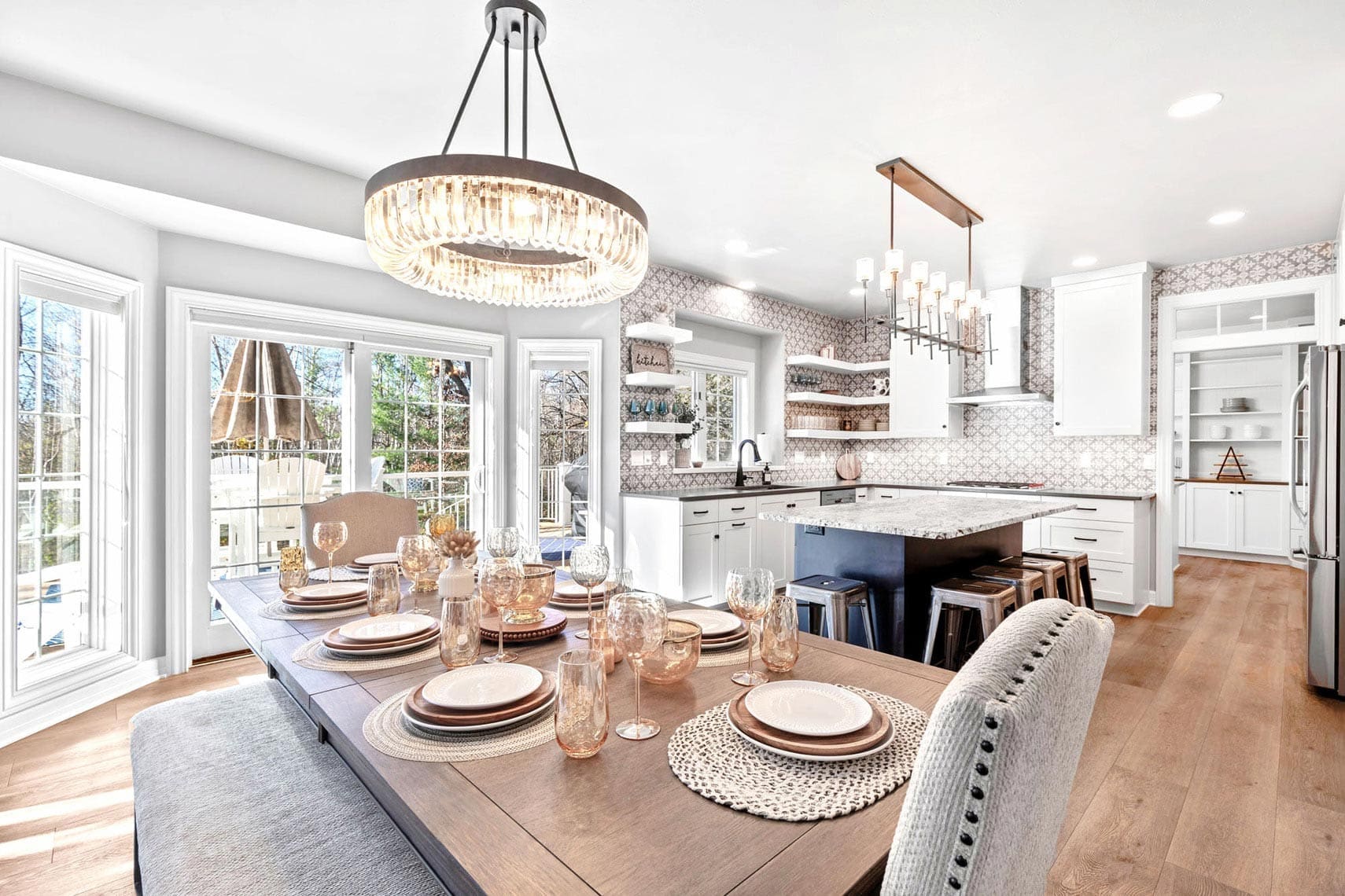 Bright kitchen and dining area in a residential property, featuring a wooden table set for eight, modern light fixtures, and white cabinets. Large windows enhance the natural lighting, showcasing the open space with light wooden flooring, ideal for real estate listings.