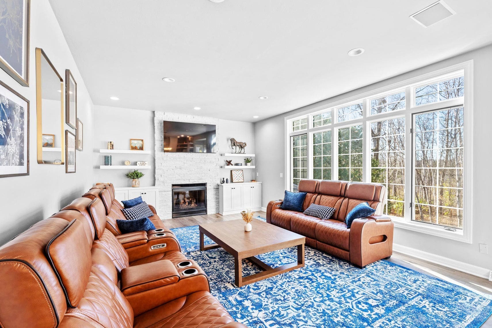 Residential real estate living room showcasing brown leather recliners, a blue patterned rug, and large windows. Features a fireplace with TV above it on the left wall and framed pictures on the opposite wall.