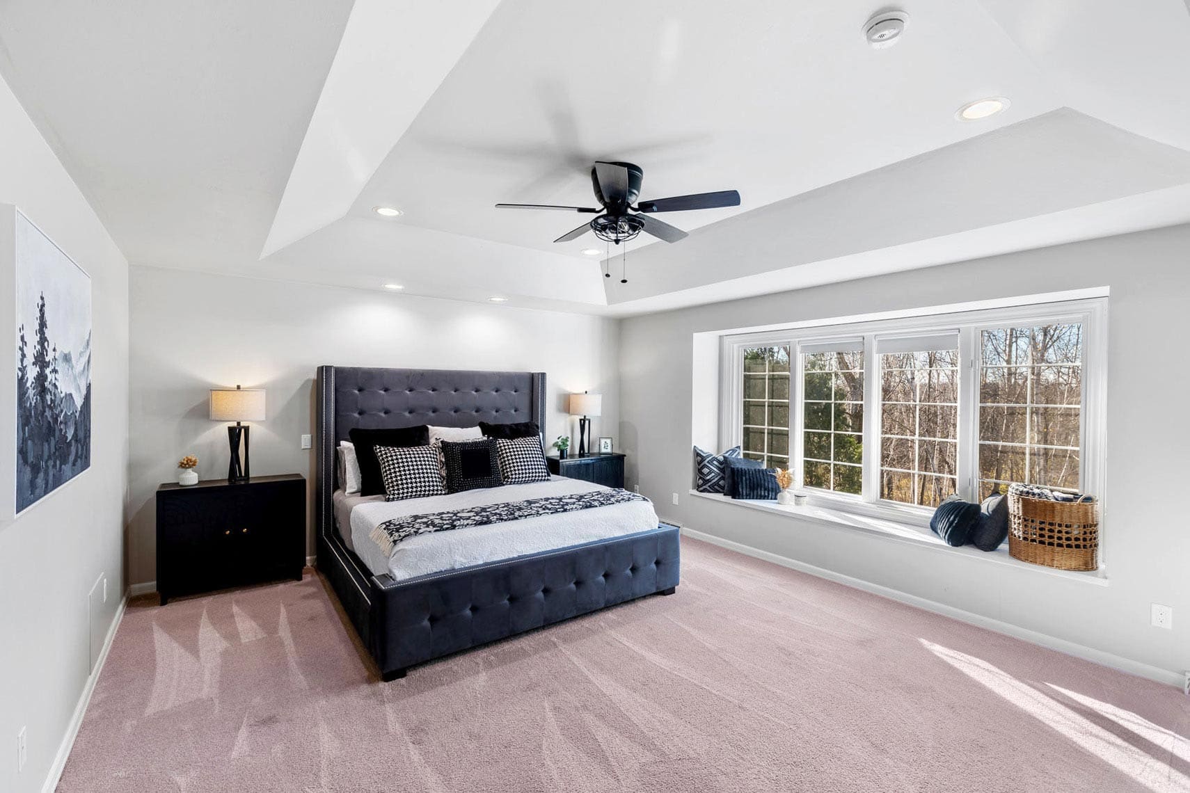 Spacious modern bedroom in residential property featuring dark upholstered bed, two bedside tables with lamps, ceiling fan, and large window seat offering outdoor views; neutral tones and minimal decor ideal for real estate listings.