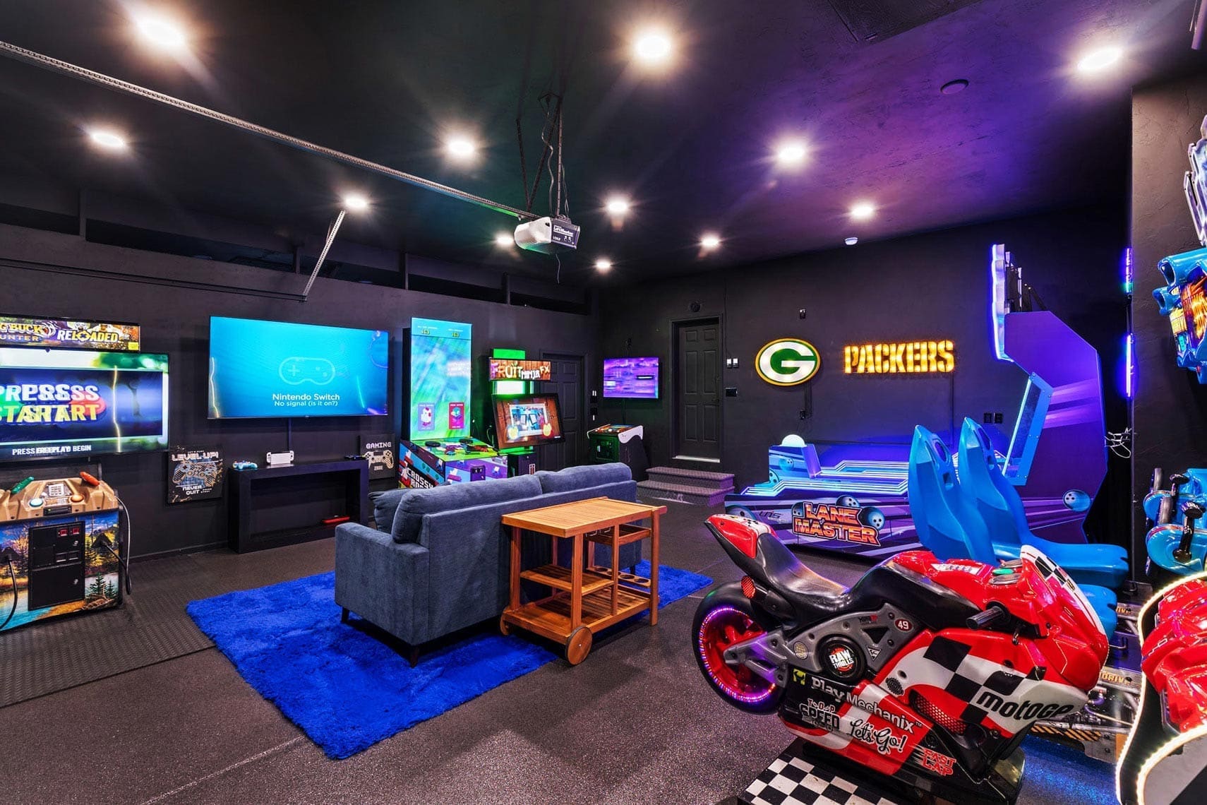 Modern gaming room in a residential property featuring arcade machines, blue sofa, bright neon lights, Packers logo, large screen TV, and red motorcycle-themed game for real estate photography.