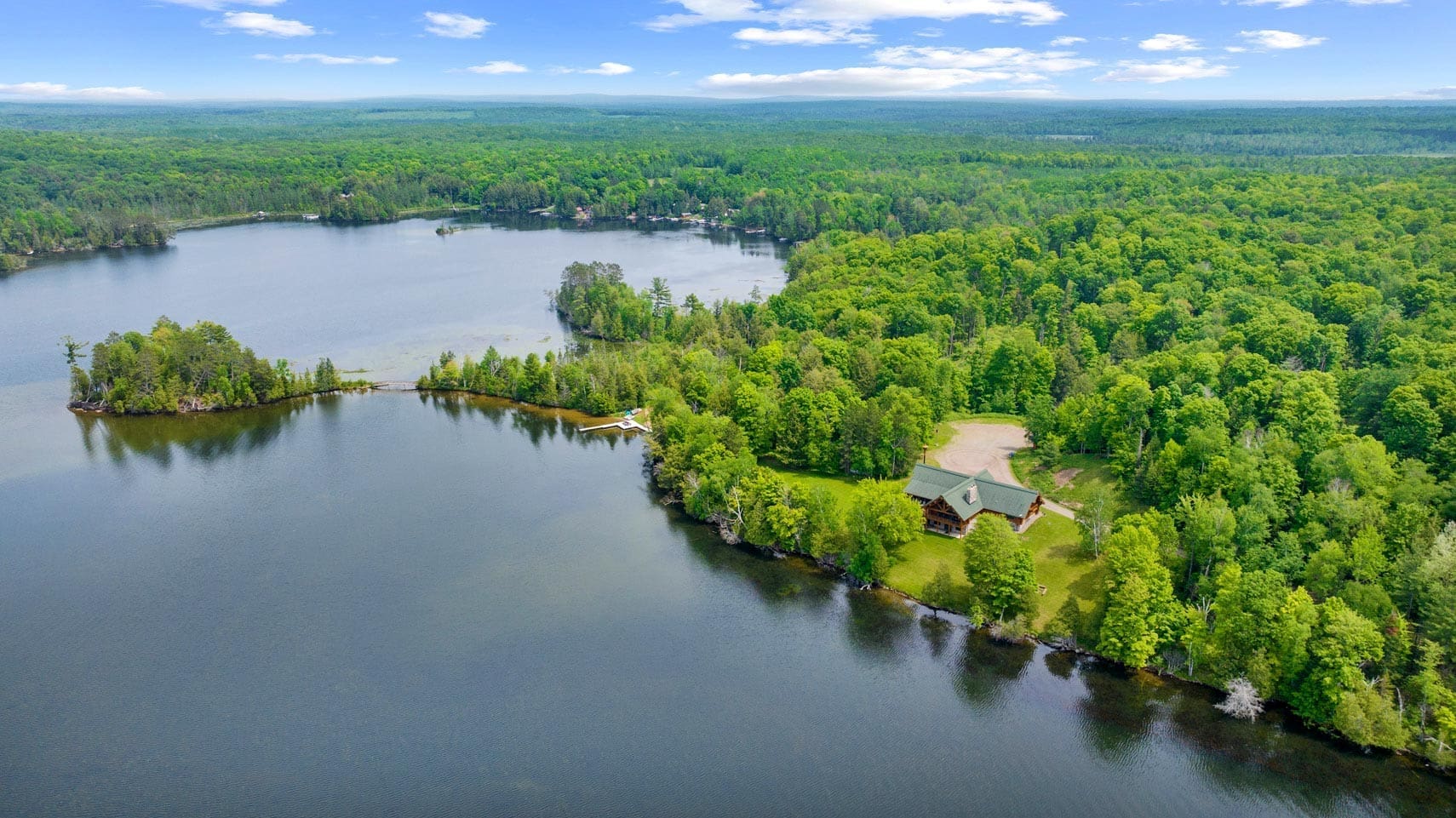 Aerial view of a picturesque lakefront property with a cozy cabin surrounded by lush green forest, ideal for residential or vacation real estate listings, under a vibrant blue sky.
