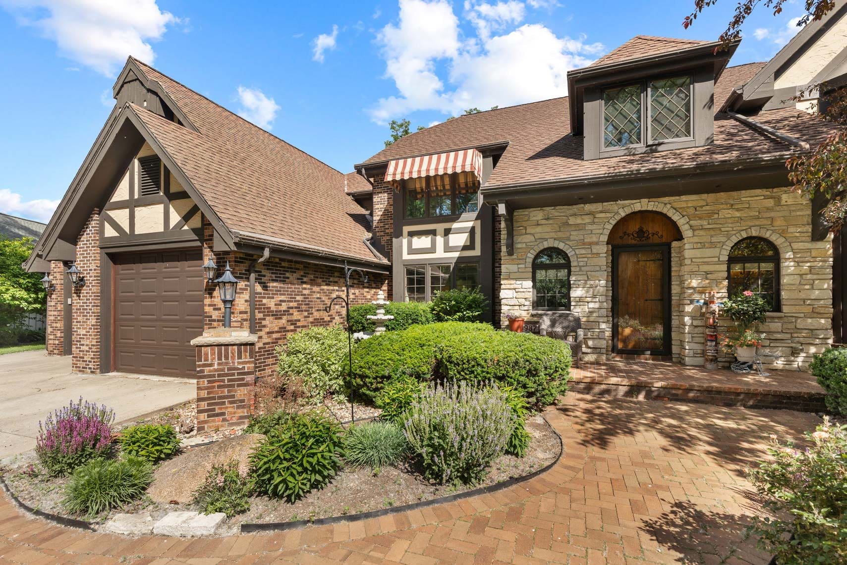 Exterior real estate photography of a brick and stone residential property featuring a brown roof, garage, and arched doorway. The home is surrounded by landscaped shrubs, flowers, and a brick path under clear blue skies, ideal for real estate marketing listings.