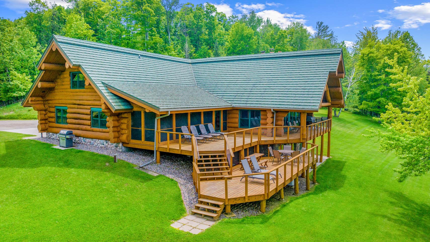 Residential log cabin with a green roof enveloped by lush trees, showcasing a wooden deck with chairs and stairs descending to a manicured lawn under a clear blue sky, ideal for real estate photography.