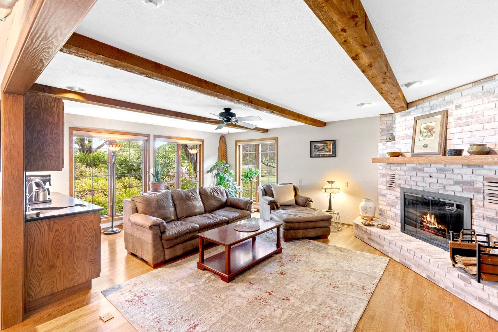 Residential real estate living room with wooden beams, large window, brown sofas, brick fireplace, glass coffee table on light rug; features ceiling fan and decorative items.