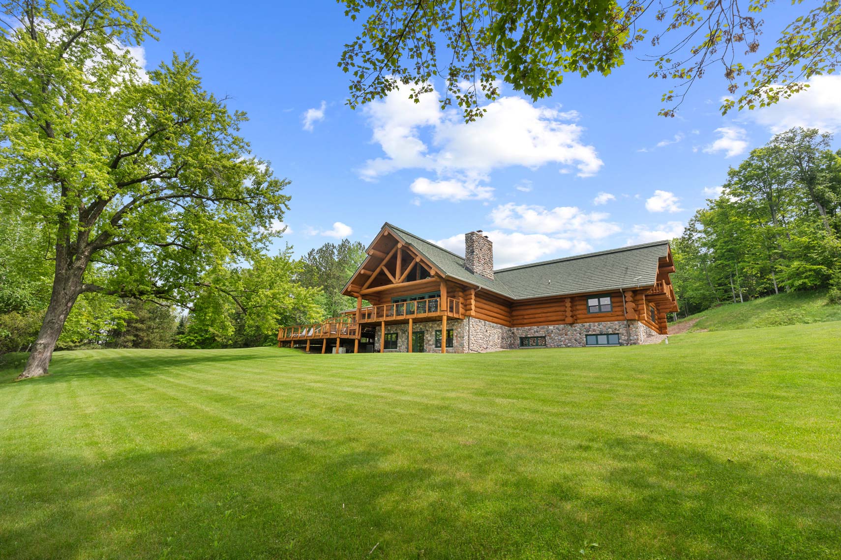 Stunning residential log cabin with stone chimney on lush green lawn, ideal for real estate listing; surrounded by trees and blue sky.