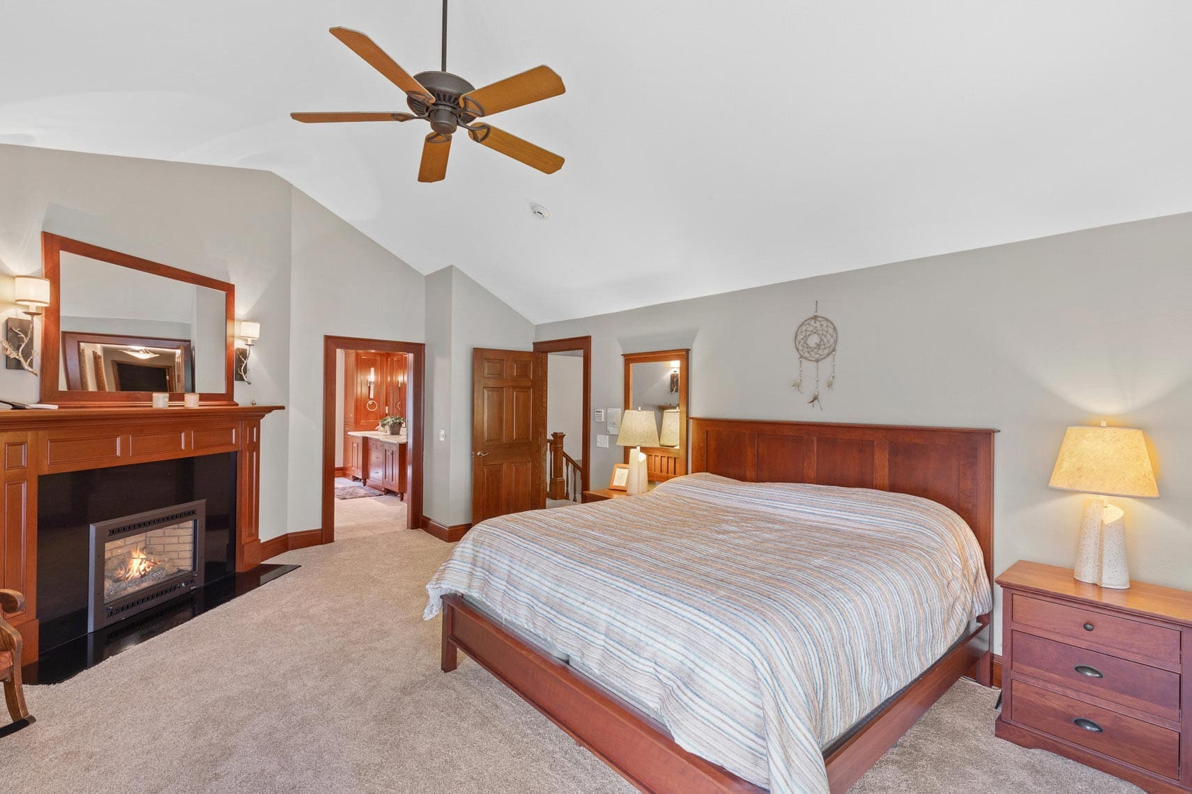 Spacious residential bedroom with a wooden bed, striped bedding, nightstands, lamps, ceiling fan, fireplace, and framed mirror. Dreamcatcher on the wall adds charm. Doorway leads to additional living space. Ideal for real estate showcasing or vacation rental listings.