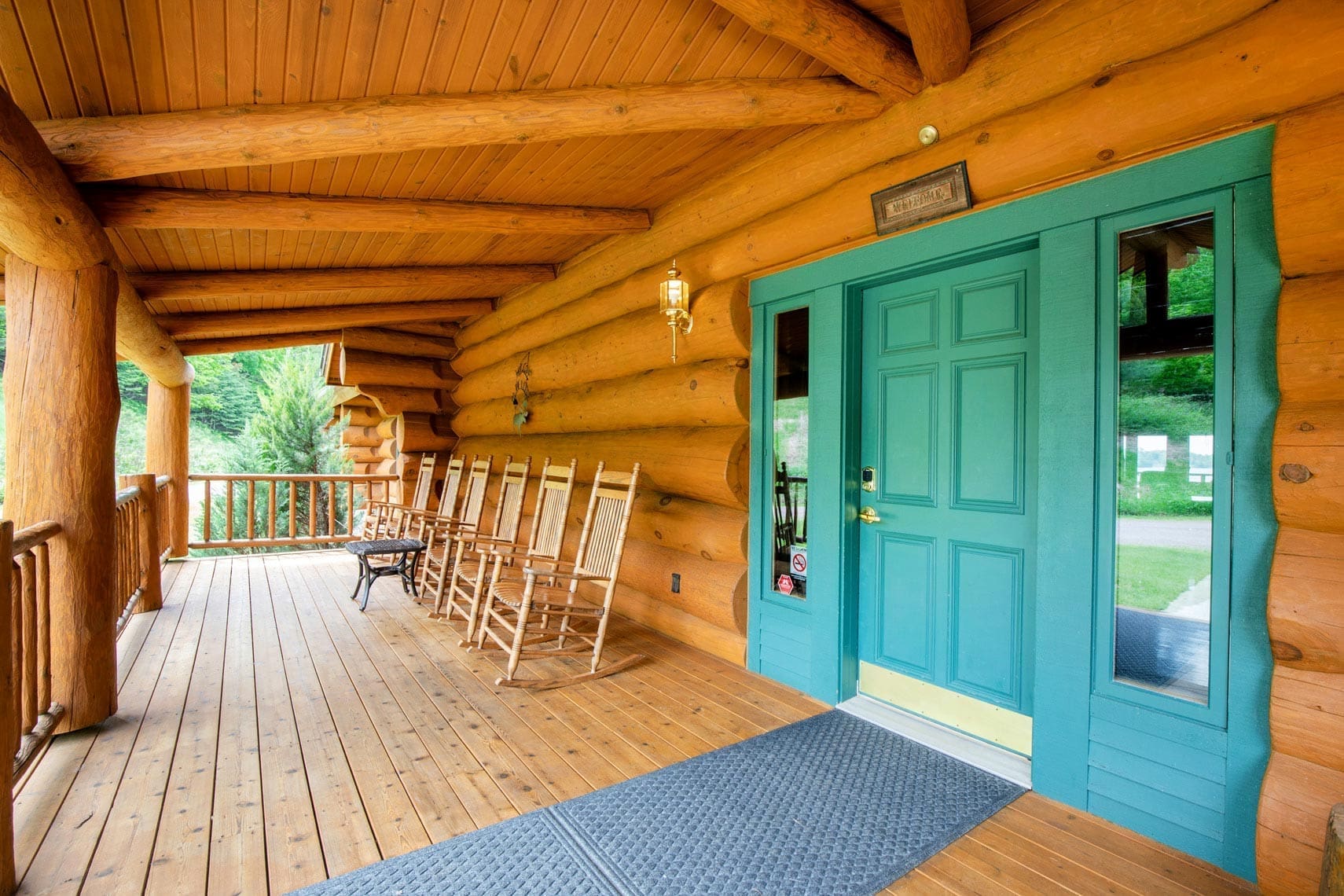 Real estate photo of a rustic log cabin porch in a residential property, featuring a blue door, several wooden rocking chairs, a lantern, and a wooden bench against the timber railing.