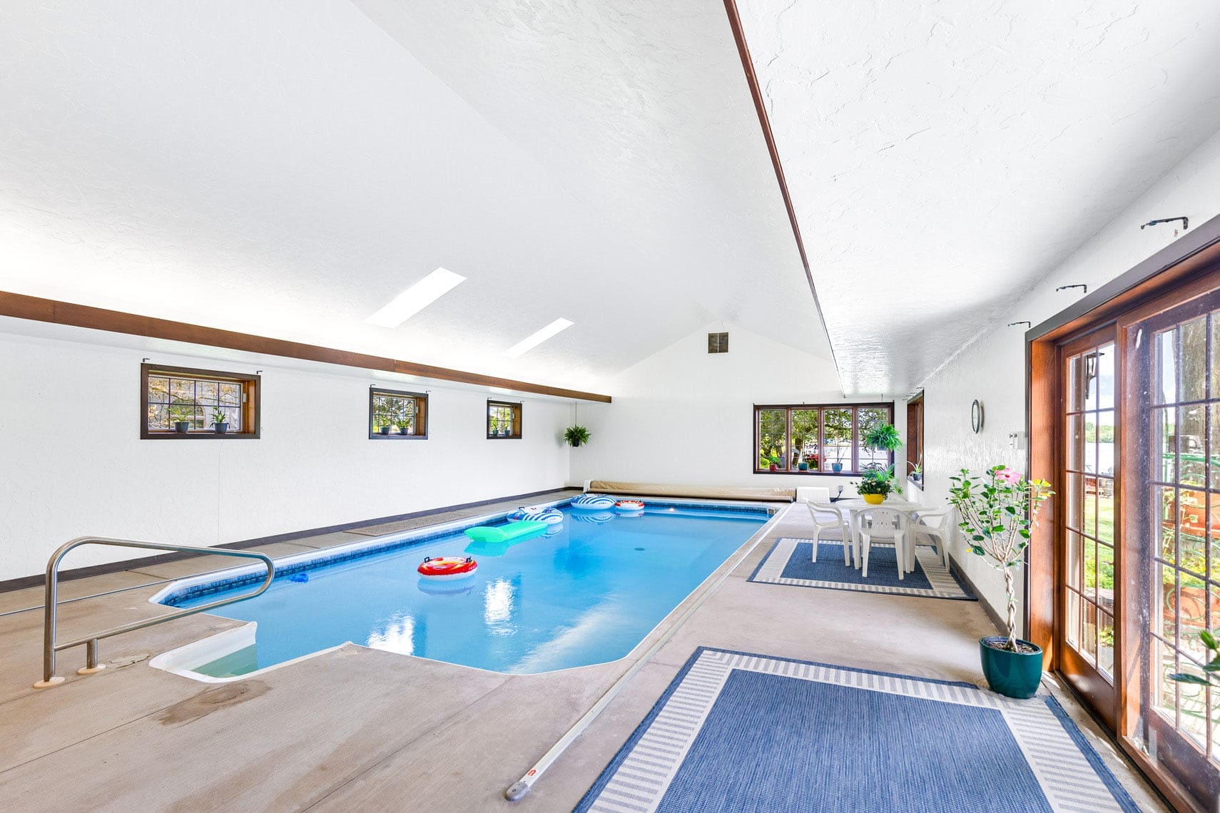Indoor swimming pool in a luxury residential property, featuring inflatables and a seating area with tables and chairs, surrounded by large windows and doors leading to an outdoor space.