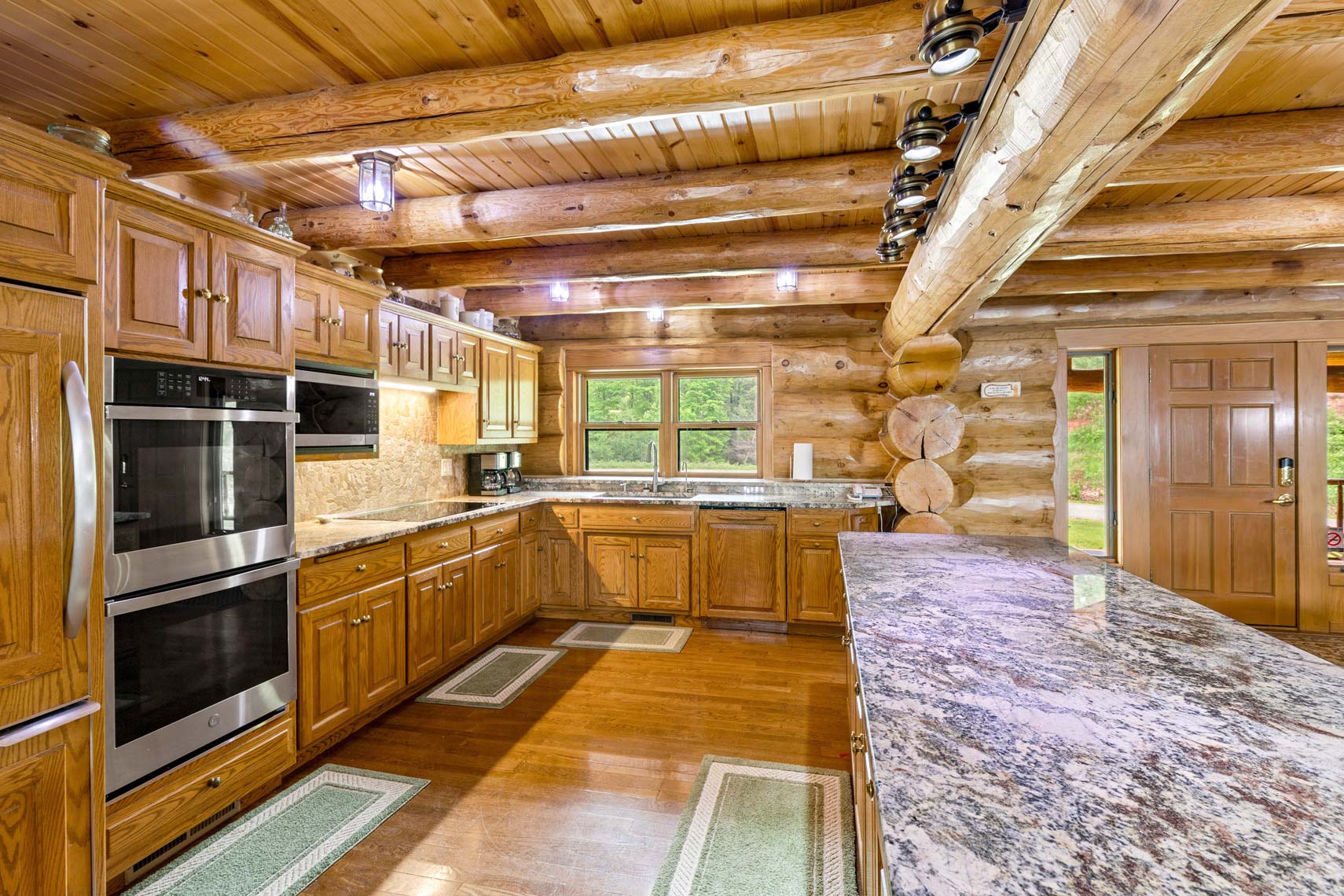 Commercial real estate photo of a spacious wooden kitchen featuring log walls, stainless steel appliances, wooden cabinets, and a granite countertop. Large window illuminates the residential space with natural light.