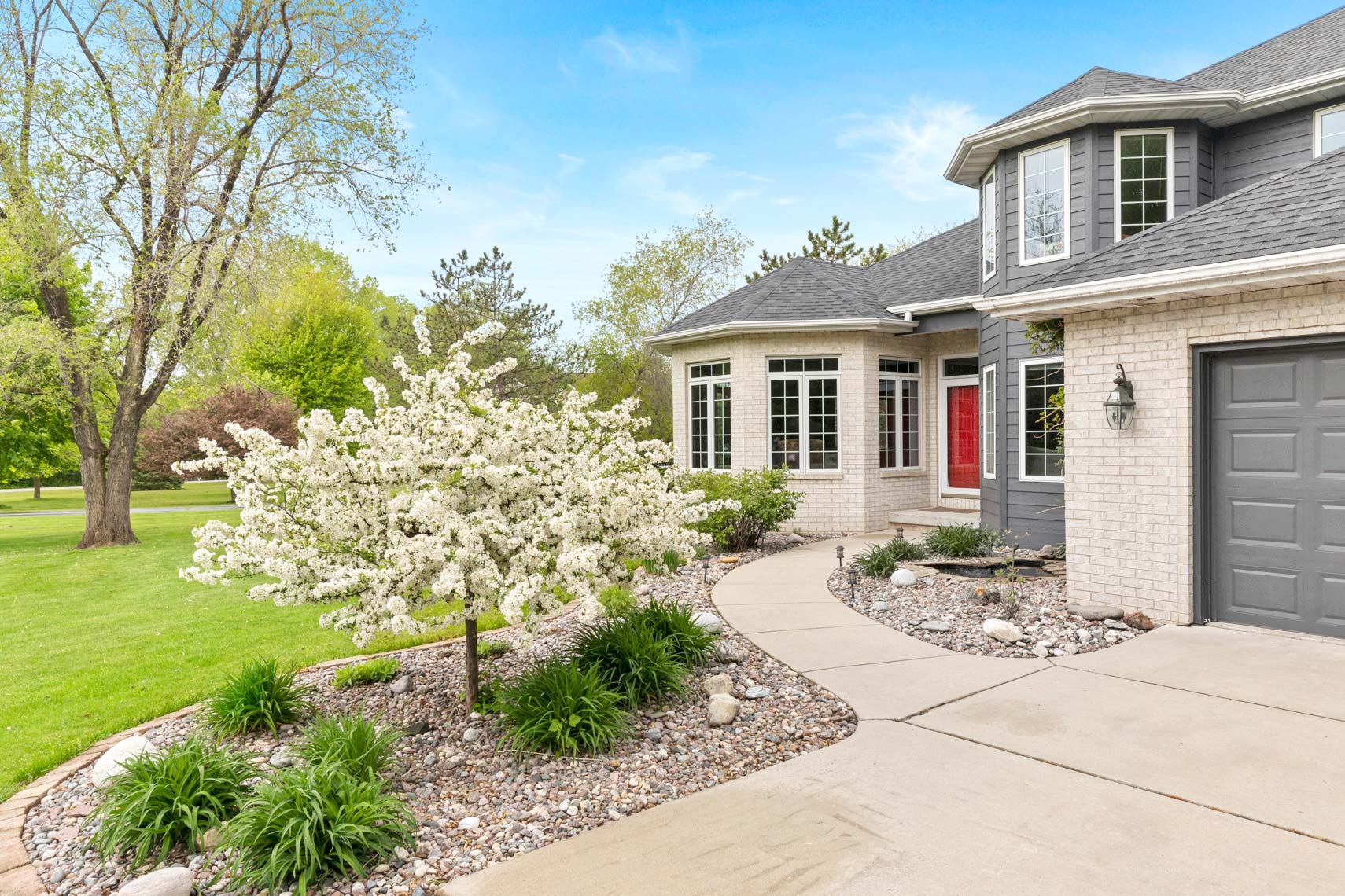 Modern residential property exterior showcasing gray siding and brick accents. Features include a curved pathway, white flowering tree, landscaped garden, and an inviting red front door suitable for real estate photography.