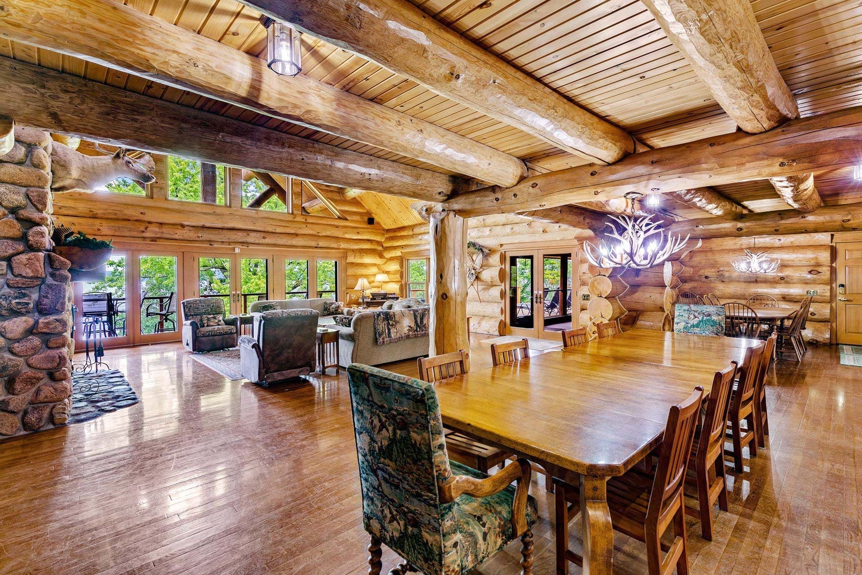 Interior of a spacious residential log cabin featuring wood paneling, a long dining table, plush seating area, large windows for natural light, and antler chandeliers—ideal for real estate photography showcasing vacation rental charm.