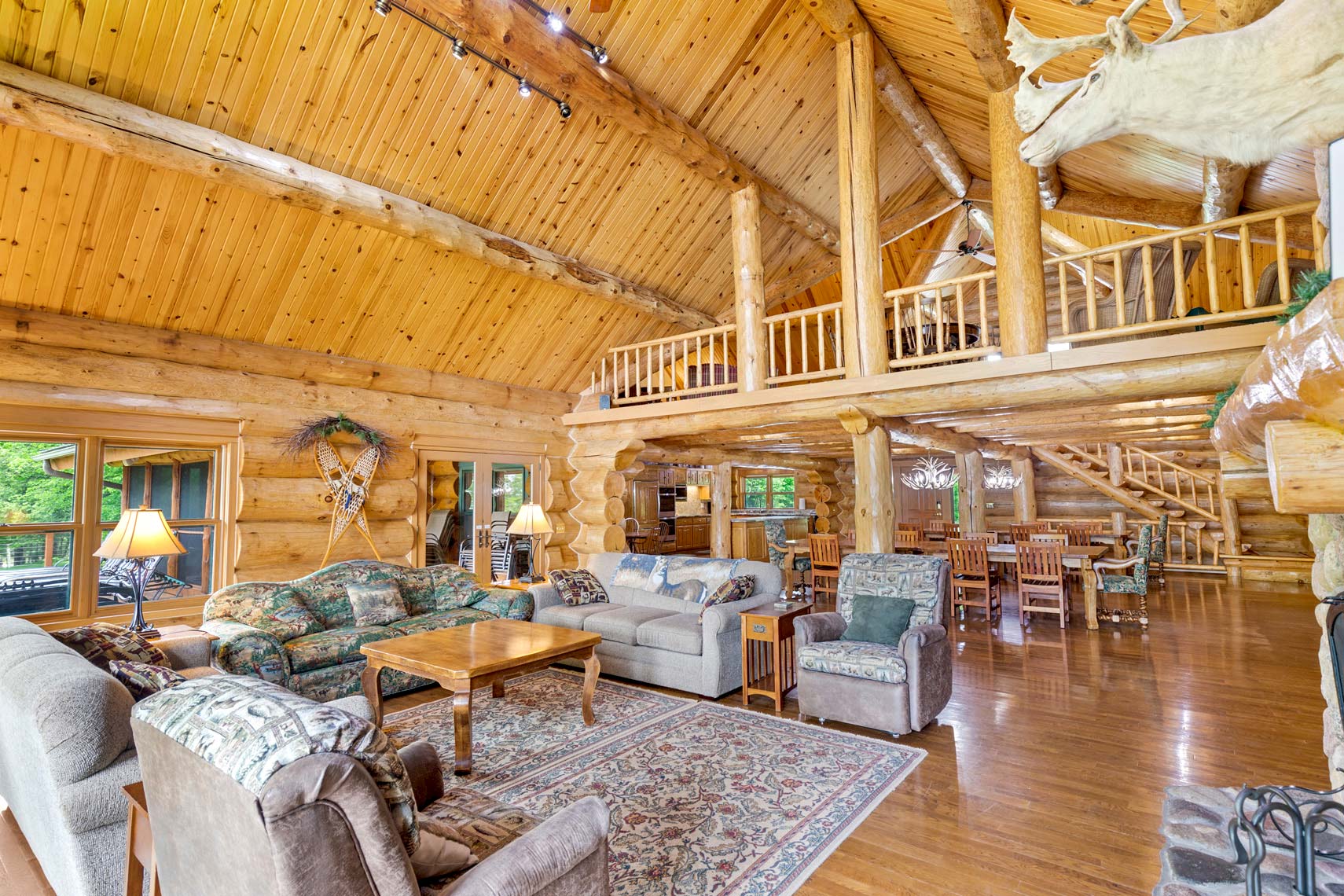 Expansive log cabin living room interior for residential property, featuring vaulted ceilings, luxurious seating, wooden dining space, and artistic mounted animal decor. Large windows and a balcony enhance natural light exposure in this real estate photography showcase.
