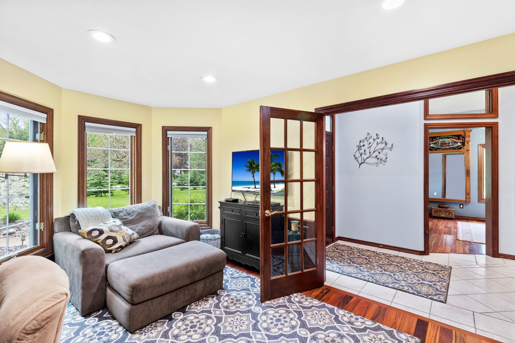 Residential real estate photo of a cozy living room featuring a large gray armchair, patterned rug, and TV displaying a beach scene. Glass doors open to another room. Includes a lamp and large windows offering views of lush greenery.