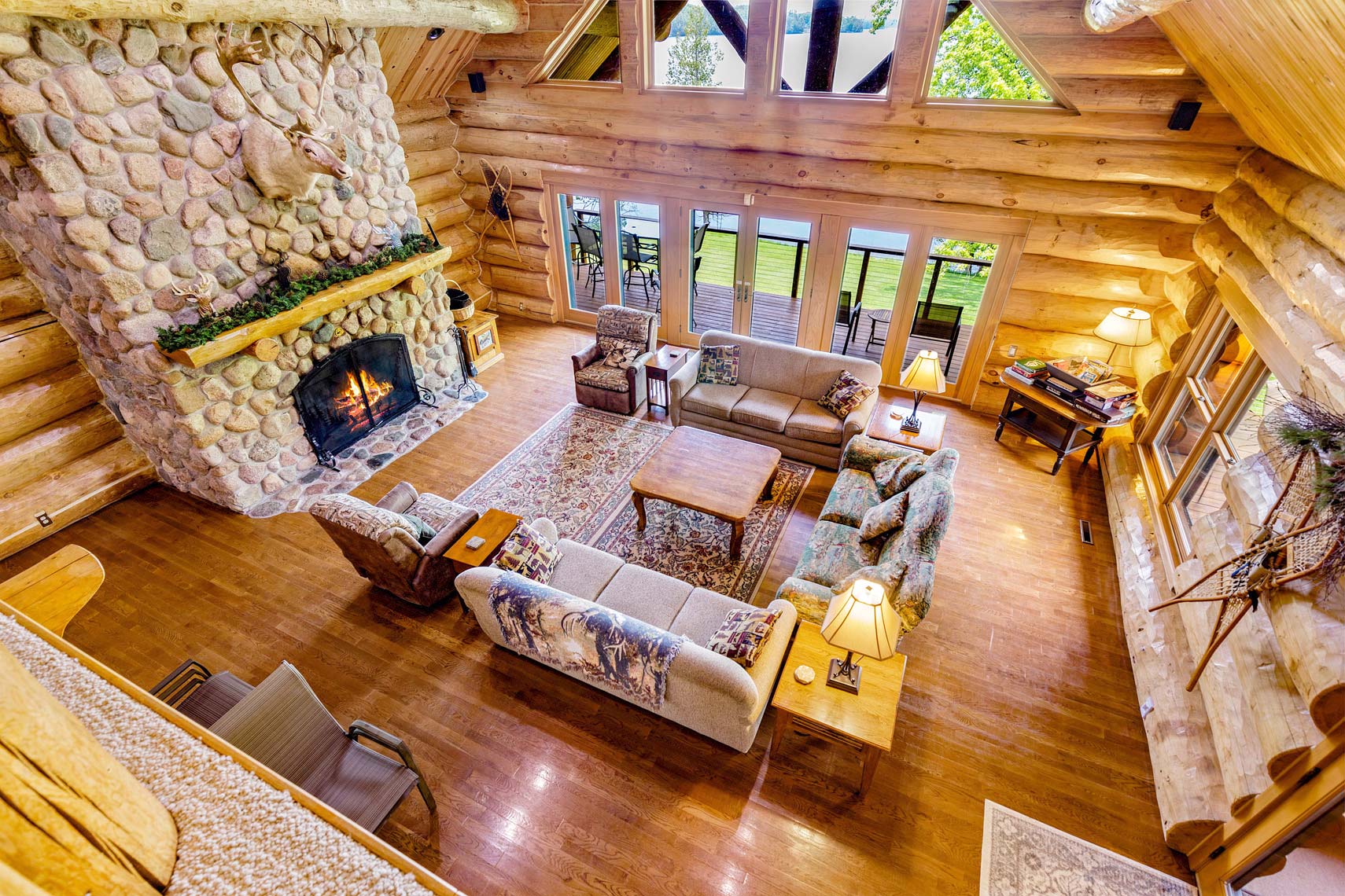 Aerial view of a cozy log cabin living room in a vacation rental property, featuring a stone fireplace, sofas, armchairs, wooden furniture, and large windows with views of the patio and surrounding greenery.