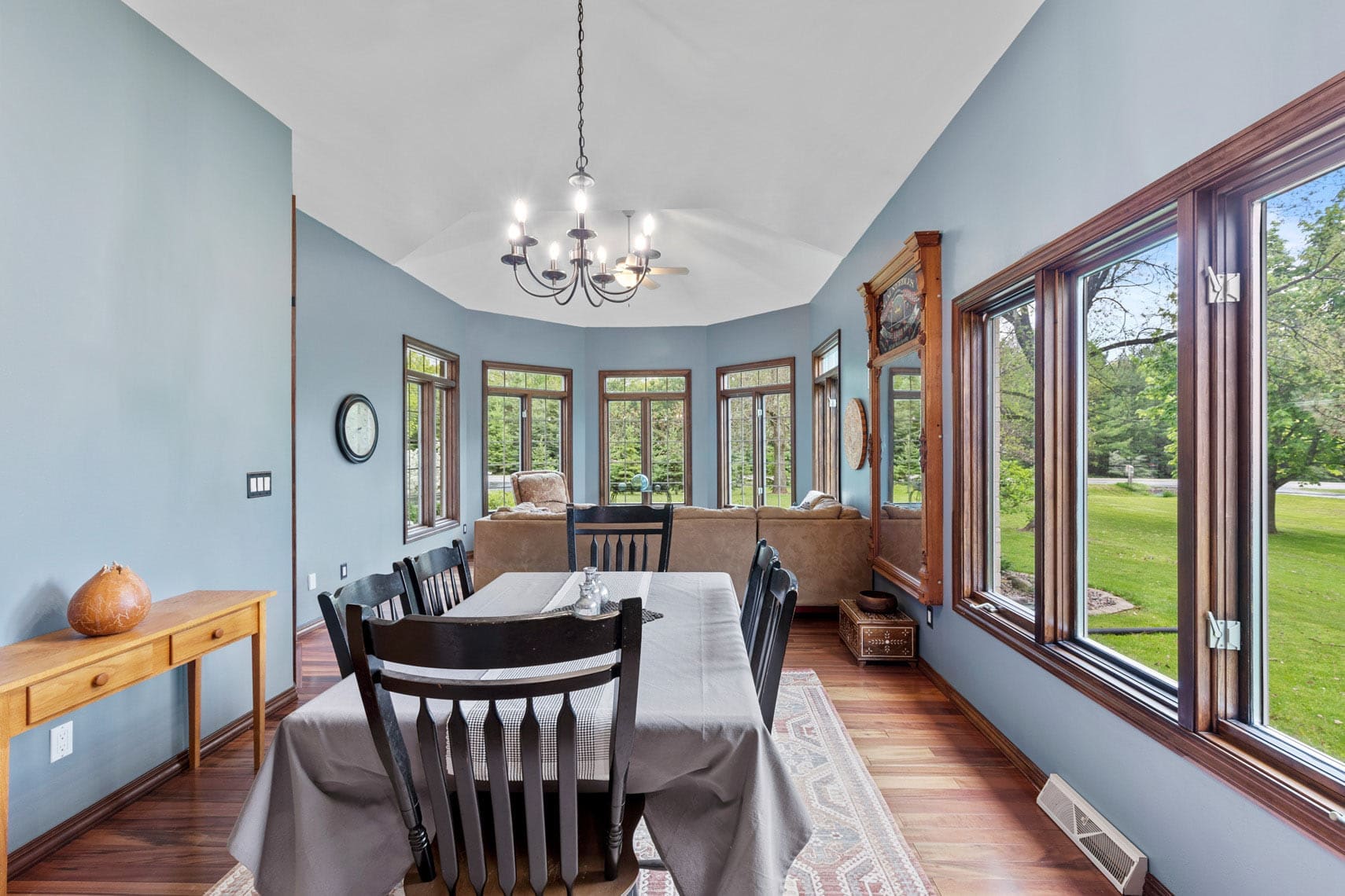 Residential real estate photo of a dining area, showcasing table and chairs next to a modern living room with a couch. Large windows highlight scenic greenery views, enhanced by chandelier lighting and elegant wooden flooring.