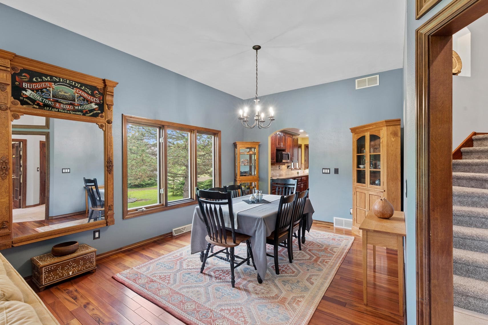 Residential real estate image showcasing a dining room with a wooden table and six chairs, featuring a chandelier above, a rug underneath, and natural light pouring in from a large window. Wood floors lead into an open-concept view of the modern kitchen and adjacent staircase.