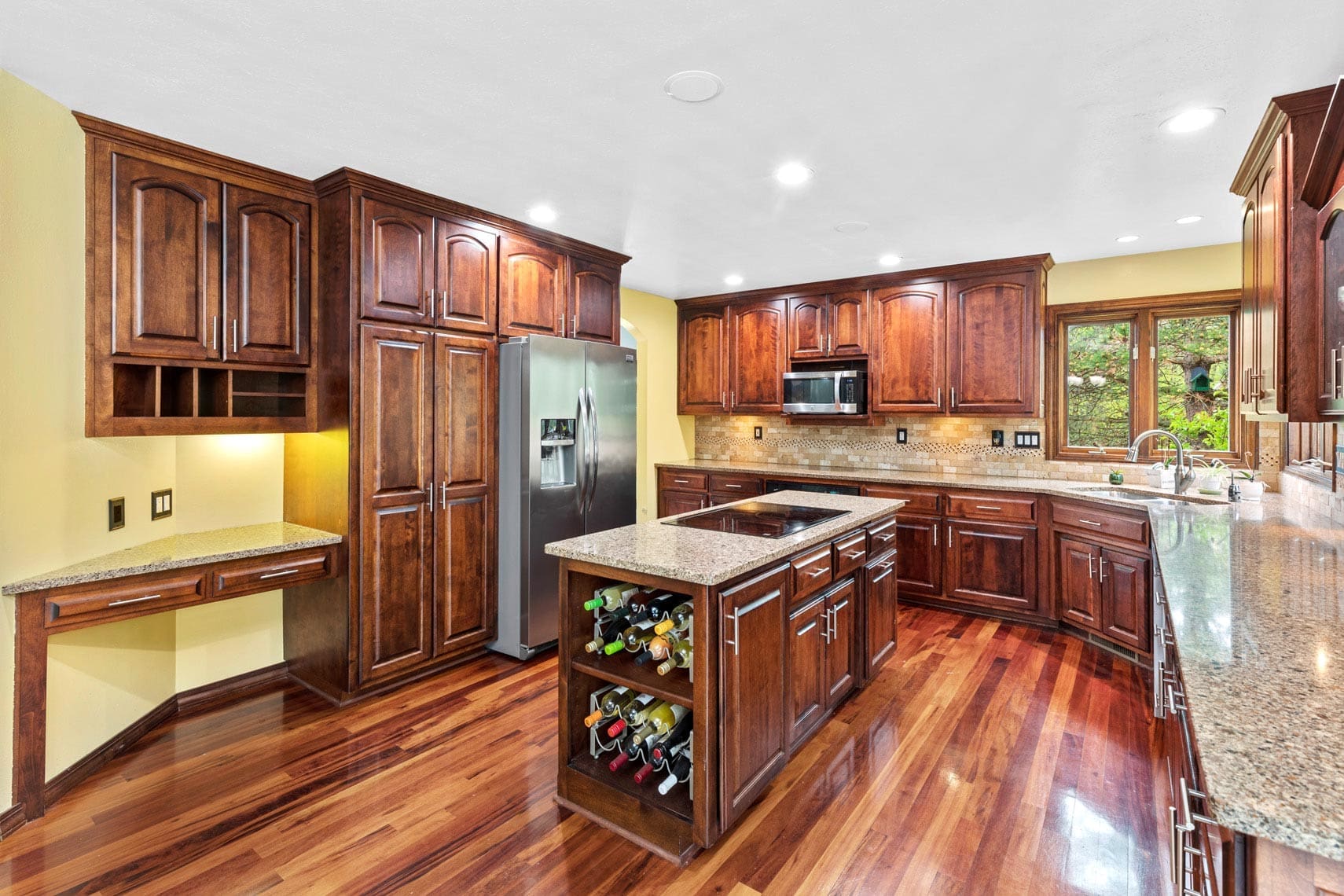Real estate photo of a spacious residential kitchen featuring wooden cabinets, granite countertops, an island with wine storage, stainless steel appliances, and hardwood floors.