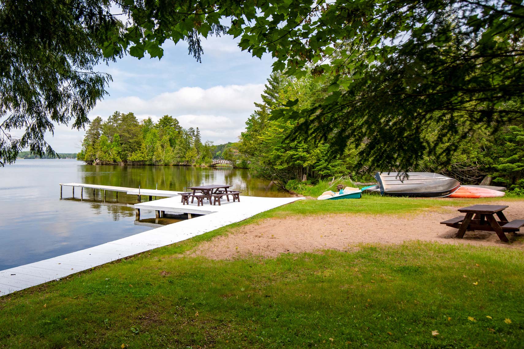 Lakeside vacation rental property with a white dock extending into the water, surrounded by lush trees. Boats and picnic tables on grassy shore under partly cloudy sky, perfect for showcasing real estate amenities.