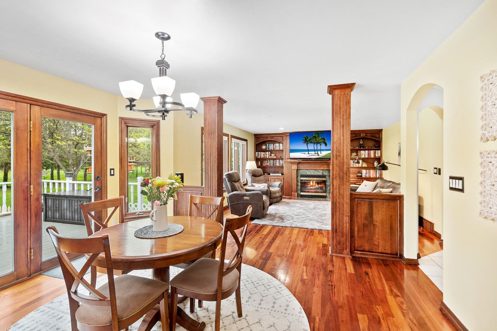 Spacious open-plan living area ideal for residential real estate featuring a wooden dining table and chairs, complemented by a cozy lounge space. Abundant natural light filters through large windows, highlighting the fireplace and mounted TV. Modern light fixtures and elegant hardwood flooring enhance the appeal of this inviting investment property or vacation rental.