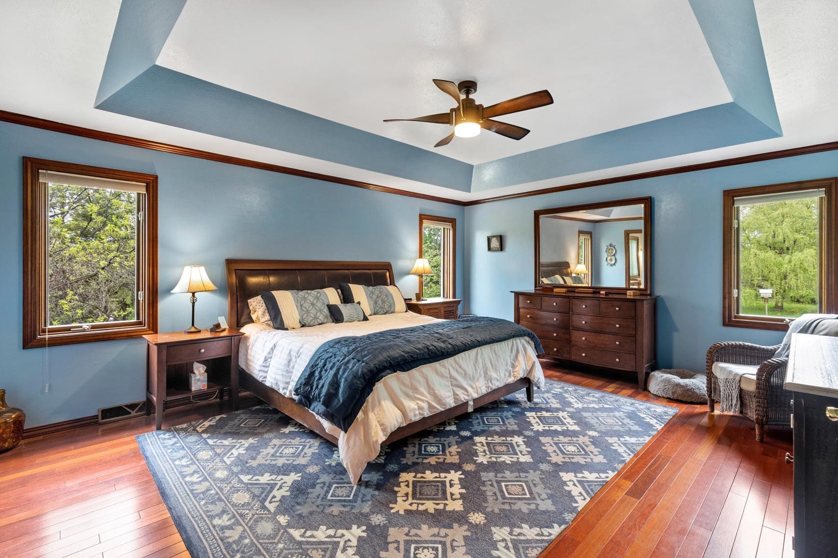 Residential bedroom showcasing blue walls and ceiling, ceiling fan, large bed with matching blue bedding, two windows for natural lighting, two decorative lamps, a dresser with mirror, and a patterned rug over polished wooden floors.