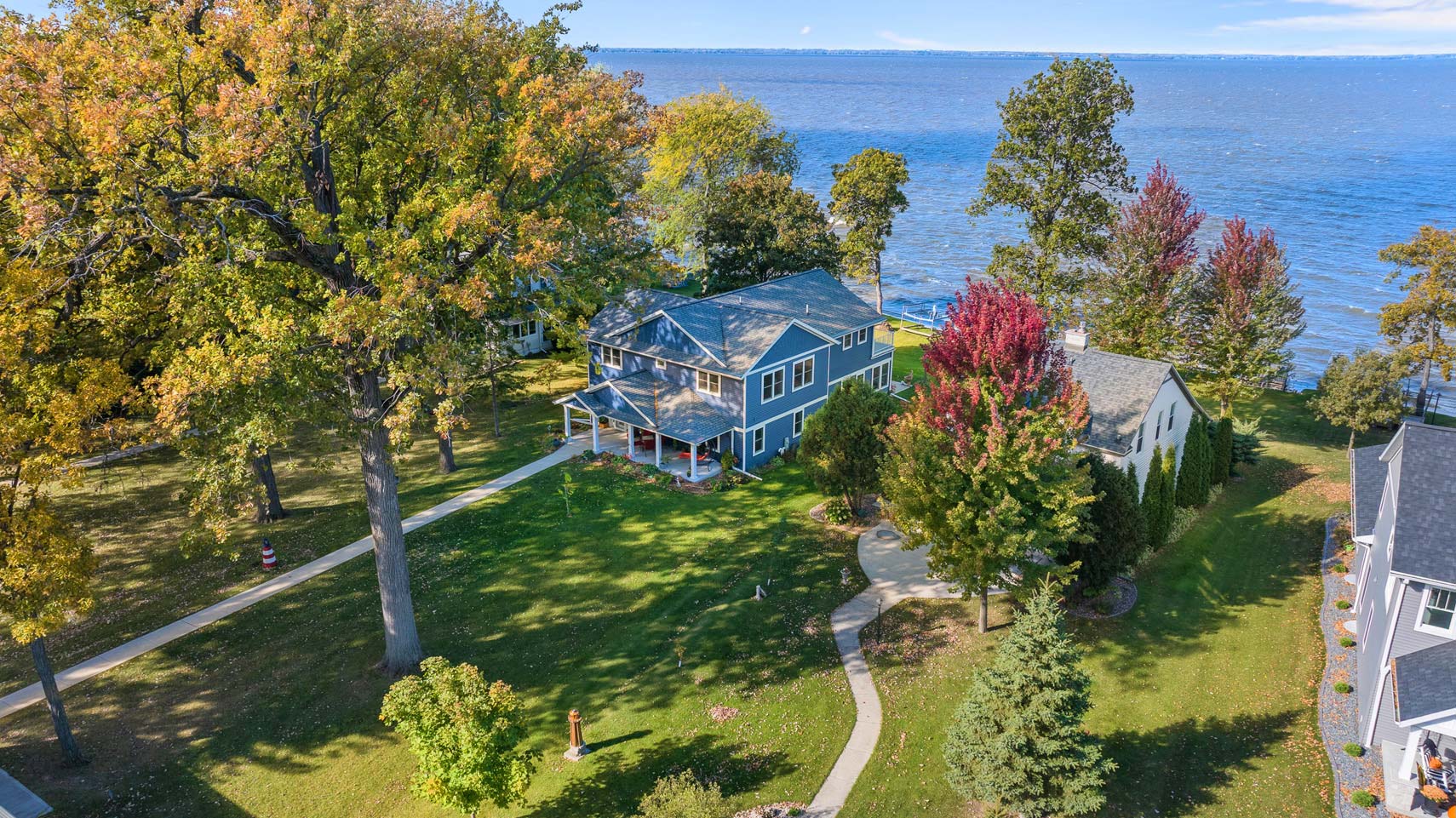 Residential real estate photography showcasing a large blue lakeside house with porch, surrounded by autumn trees and lush green yard; features a winding path leading to the lakefront.