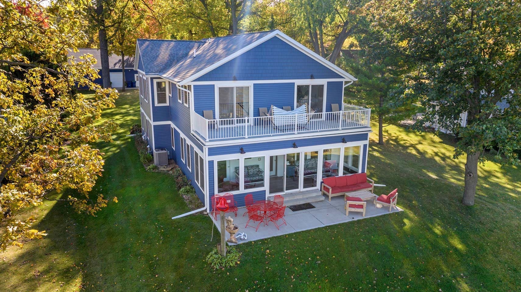 Two-story blue residential home with a white balcony, surrounded by lush trees. Features a red outdoor seating area on the patio and well-maintained landscaping, ideal for real estate listings and property showcases.