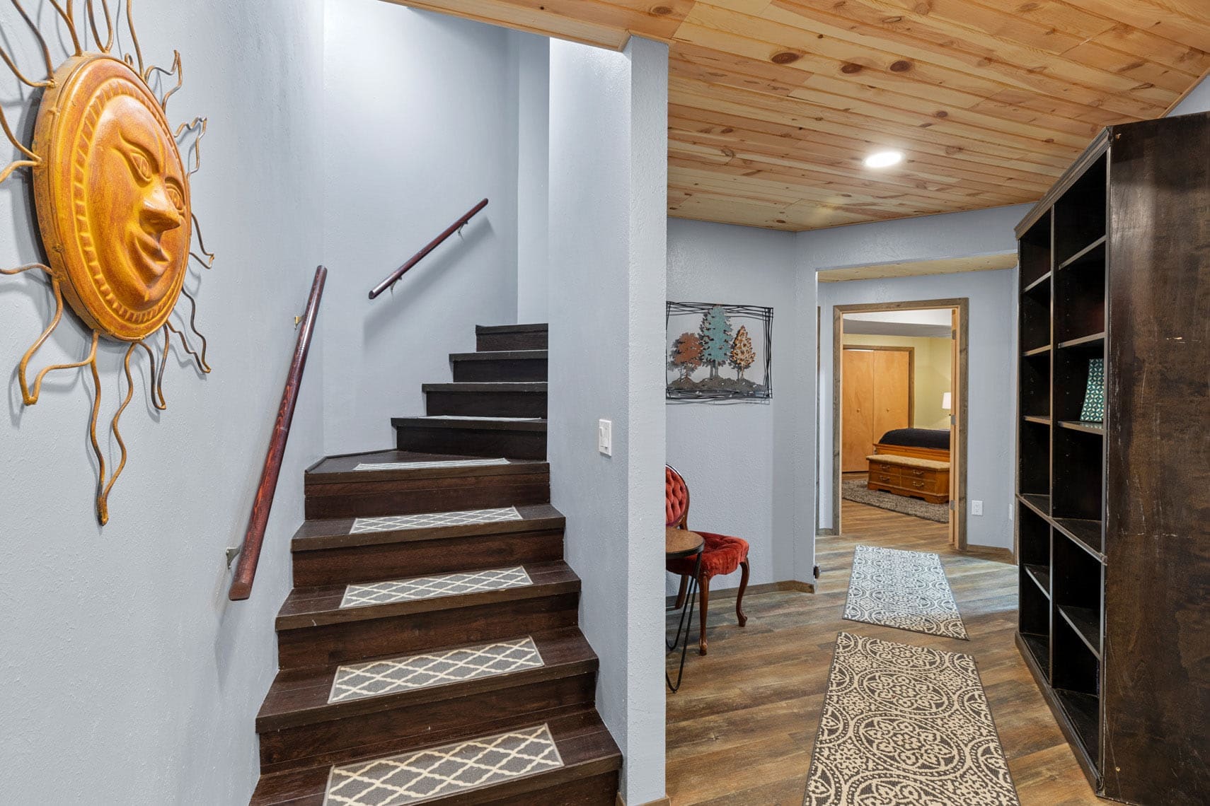 Wooden staircase in a cozy residential property interior with sun wall decoration, wooden ceiling, and patterned rugs on the hallway floor.