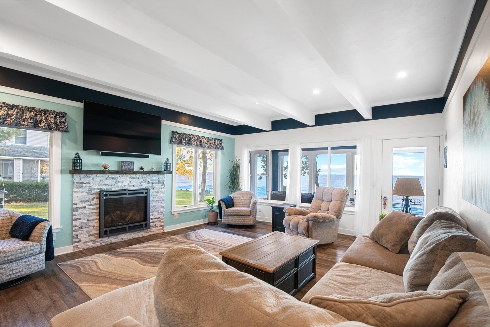 Residential real estate living room featuring beige sofas, a fireplace, and wall-mounted TV. Large windows showcase a stunning lake view. Decor includes armchairs, a coffee table, and a patterned rug ideal for real estate listings.
