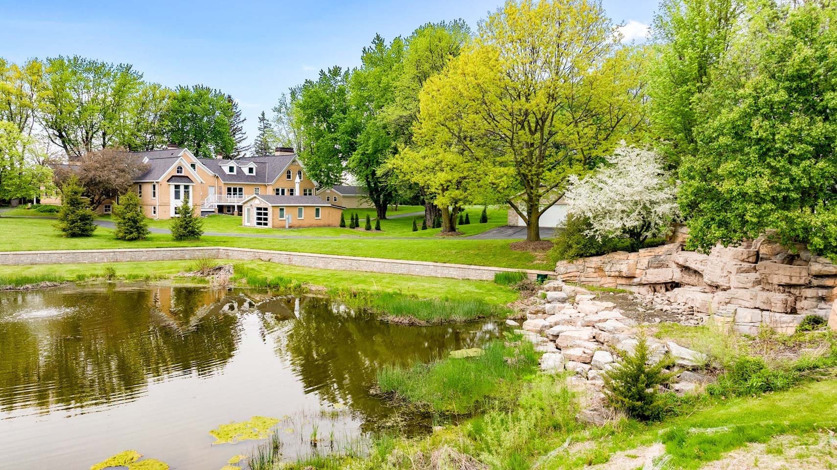 Large residential property with tan siding and multiple architectural peaks, surrounded by lush greenery, adjacent to a scenic pond with a stone wall and mature trees in the background. Ideal for real estate photography showcasing tranquil living environments.