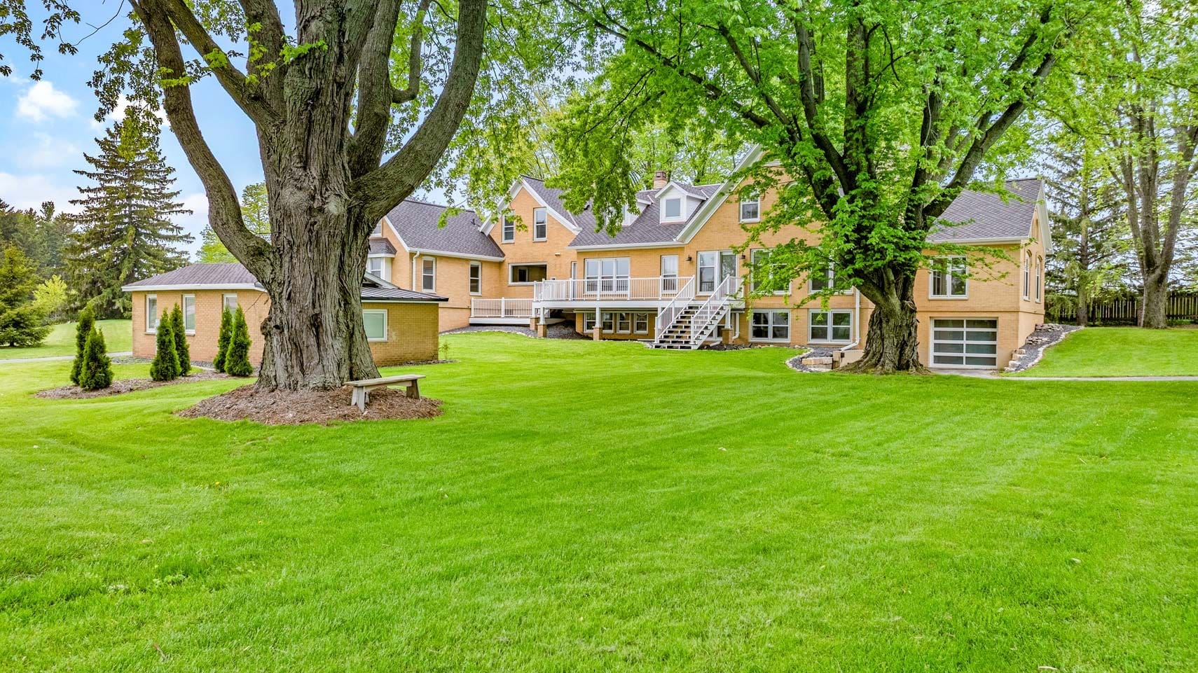 A two-story brick residential property with multiple gables, surrounded by lush trees and a meticulously maintained lawn, showcasing a wooden deck and stone path, perfect for real estate listings under a clear blue sky.