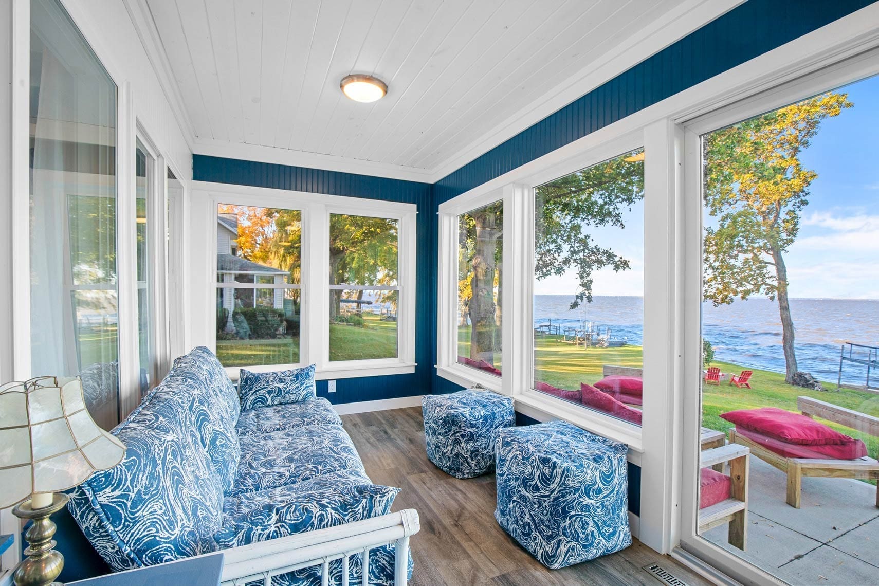 Residential real estate sunroom with blue floral furniture, large windows showcasing lake view, and outdoor patio featuring red chairs and firepit.