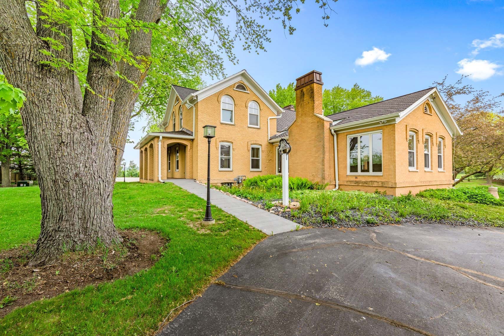 Residential real estate photo of a charming brick house with a gabled roof, surrounded by mature trees and a manicured lawn, featuring a paved walkway leading to the welcoming entrance. A decorative lamp post enhances the inviting curb appeal.