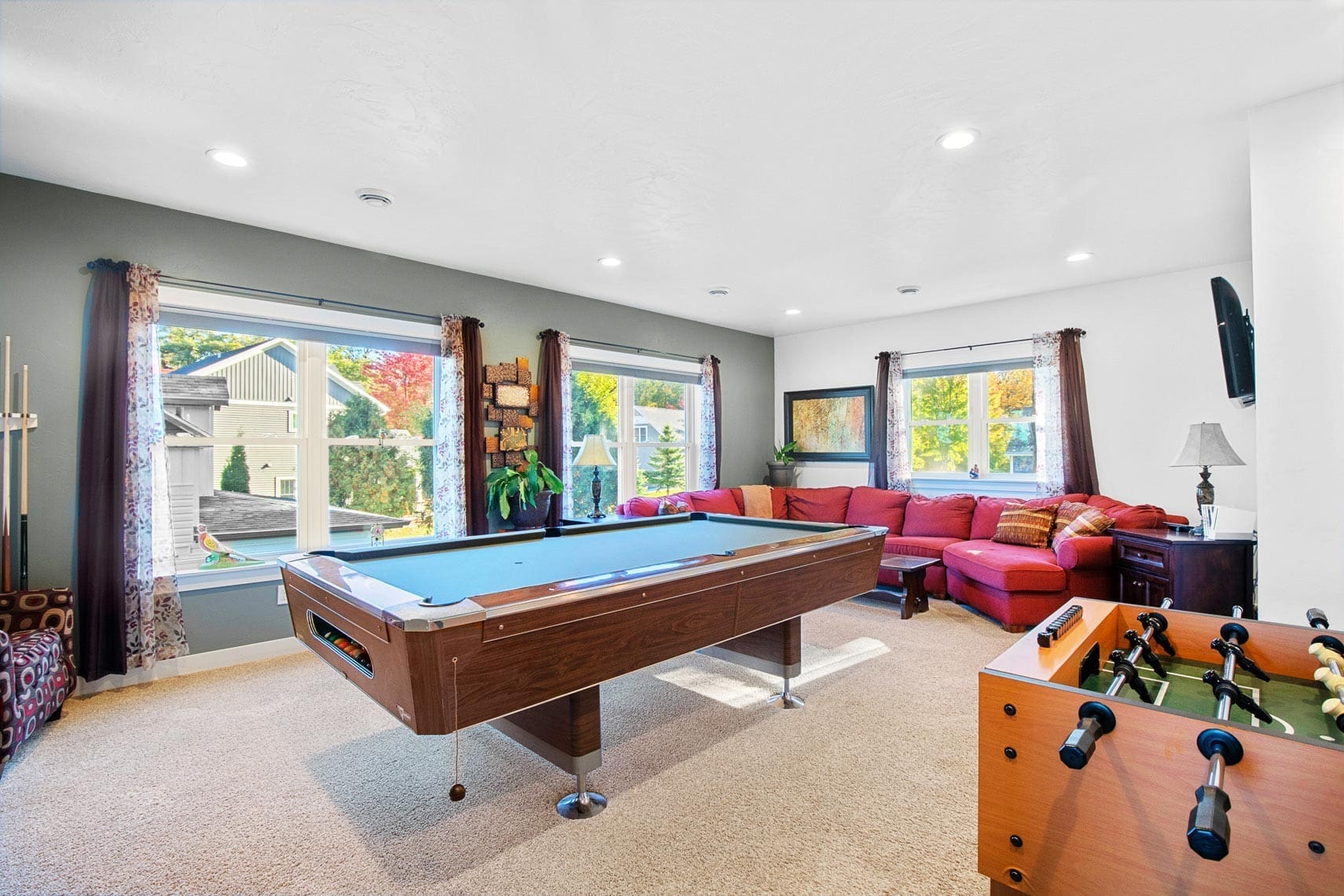 Residential entertainment room featuring a pool table, foosball table, and red sectional sofa; large windows offer natural light and scenic views of trees and nearby properties.