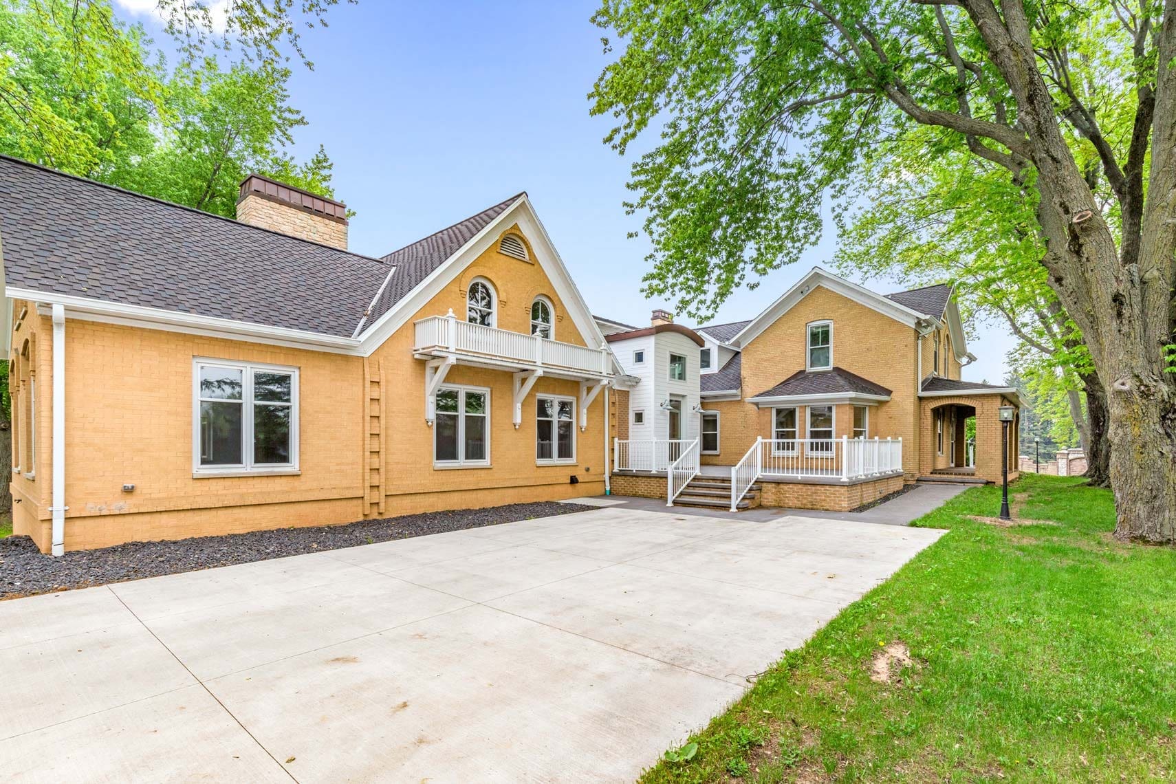 Residential real estate photography: Spacious paved driveway leading to two tan single-family homes, surrounded by mature trees and a manicured grassy area on the right.