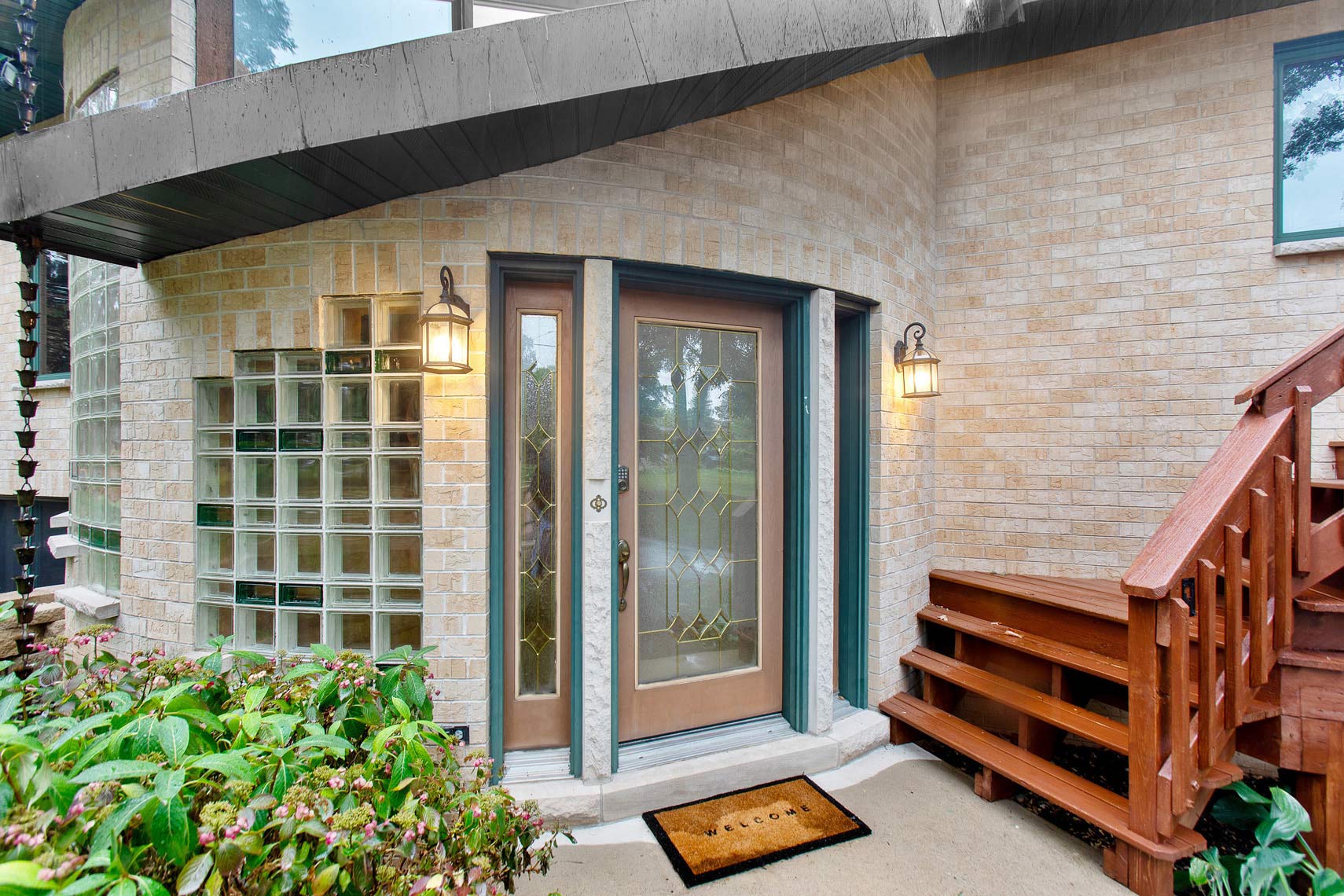 Residential property front entrance with a glass-paneled door, wall-mounted lanterns, welcome mat, wooden steps, and lush landscaping.