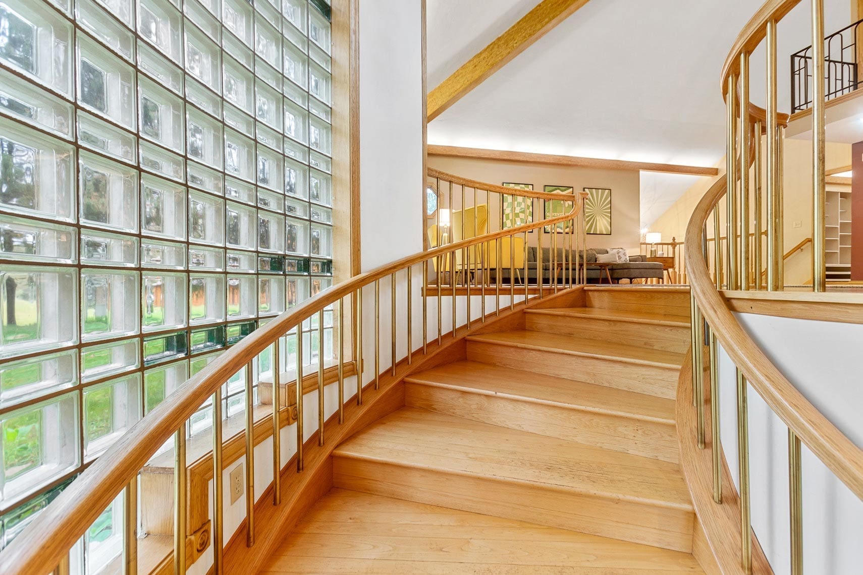 Residential property interior featuring a curved wooden staircase with metal railing, adjacent to a glass block wall, leading to an open living area with modern furniture and artwork.