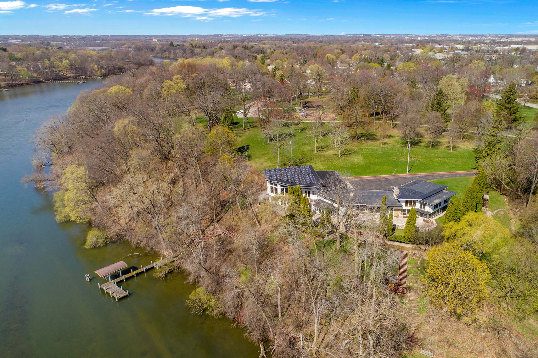Aerial view of a large residential property with solar panels, surrounded by lush trees and lakeside features, including a small dock. The scenic landscape showcases expansive green spaces and nearby real estate properties, ideal for commercial or vacation rental opportunities.