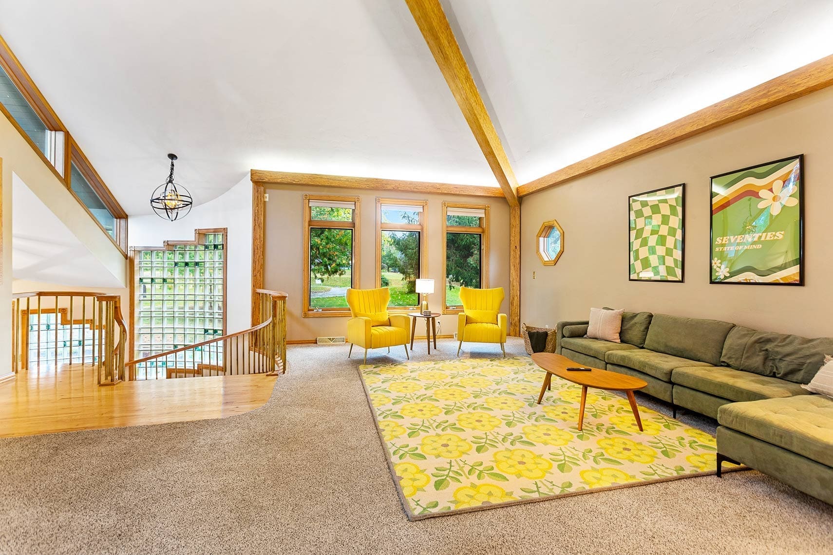 Residential real estate photography of a spacious living room featuring a green sofa, two yellow chairs, wooden coffee table, and floral rug; enhanced by artwork and large windows. Stairs with wooden rails add architectural interest.