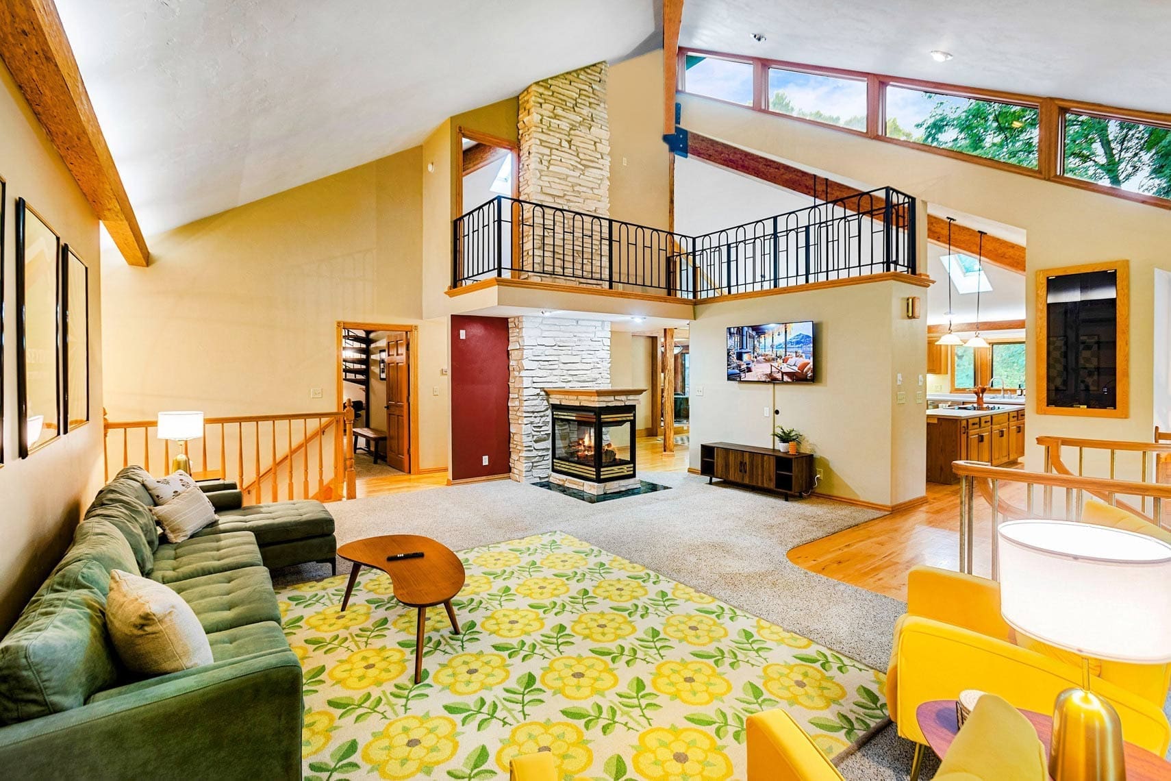 Spacious residential living room with a green sofa, yellow armchairs, a floral rug, stone fireplace, and high vaulted ceilings. Large windows offer abundant natural light for stunning real estate photography. Mezzanine visible above.