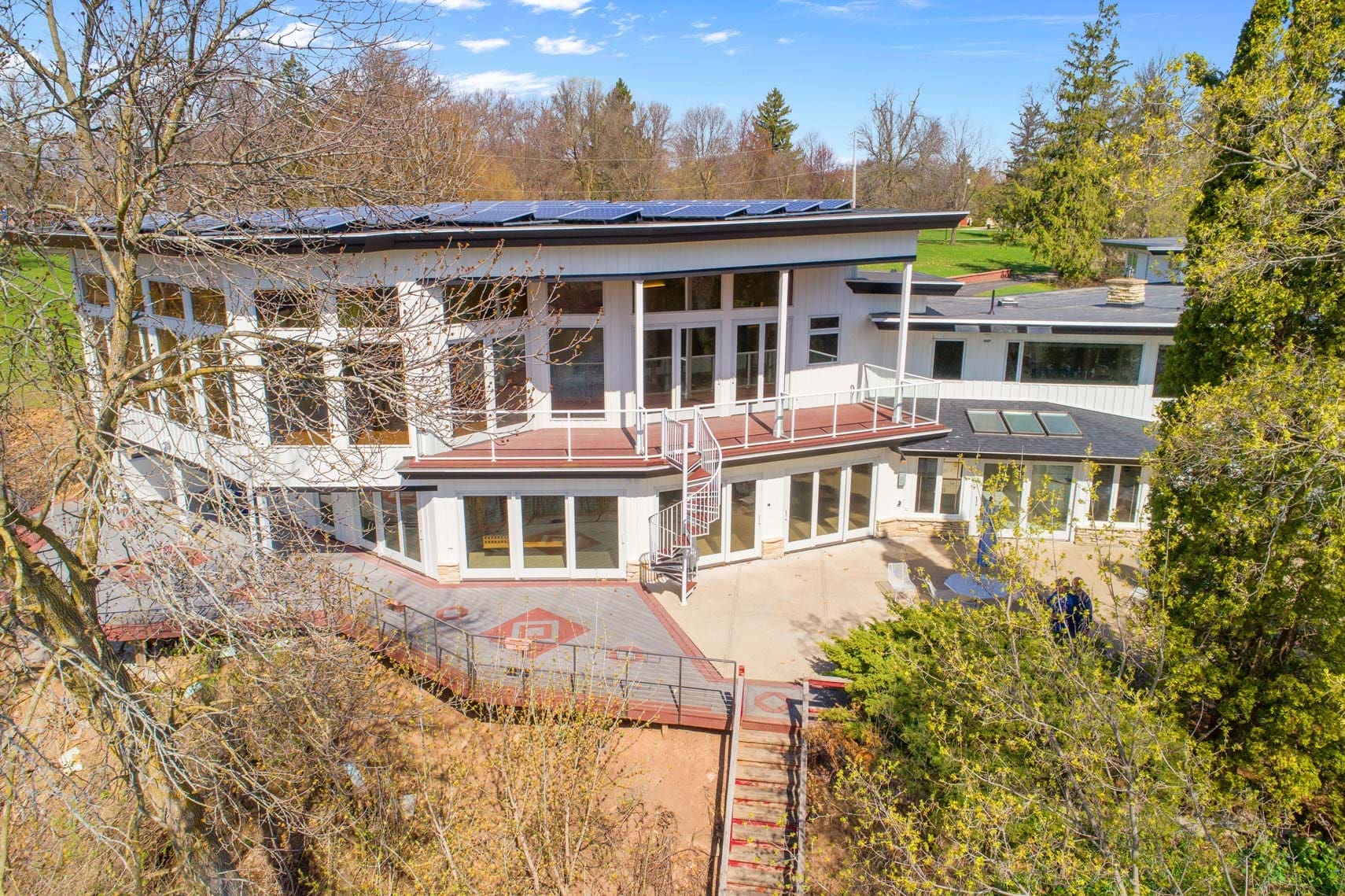 Contemporary multi-level residential property with large windows and solar panels on the roof, featuring a spacious deck. Nestled amidst trees and lush greenery, ideal for showcasing energy-efficient real estate.