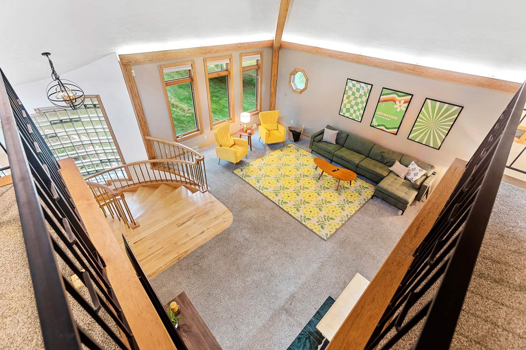 Aerial real estate photography showcasing a modern residential living room featuring a spiral staircase, yellow armchairs, a green sofa, and a patterned rug. Large windows offer abundant natural light.