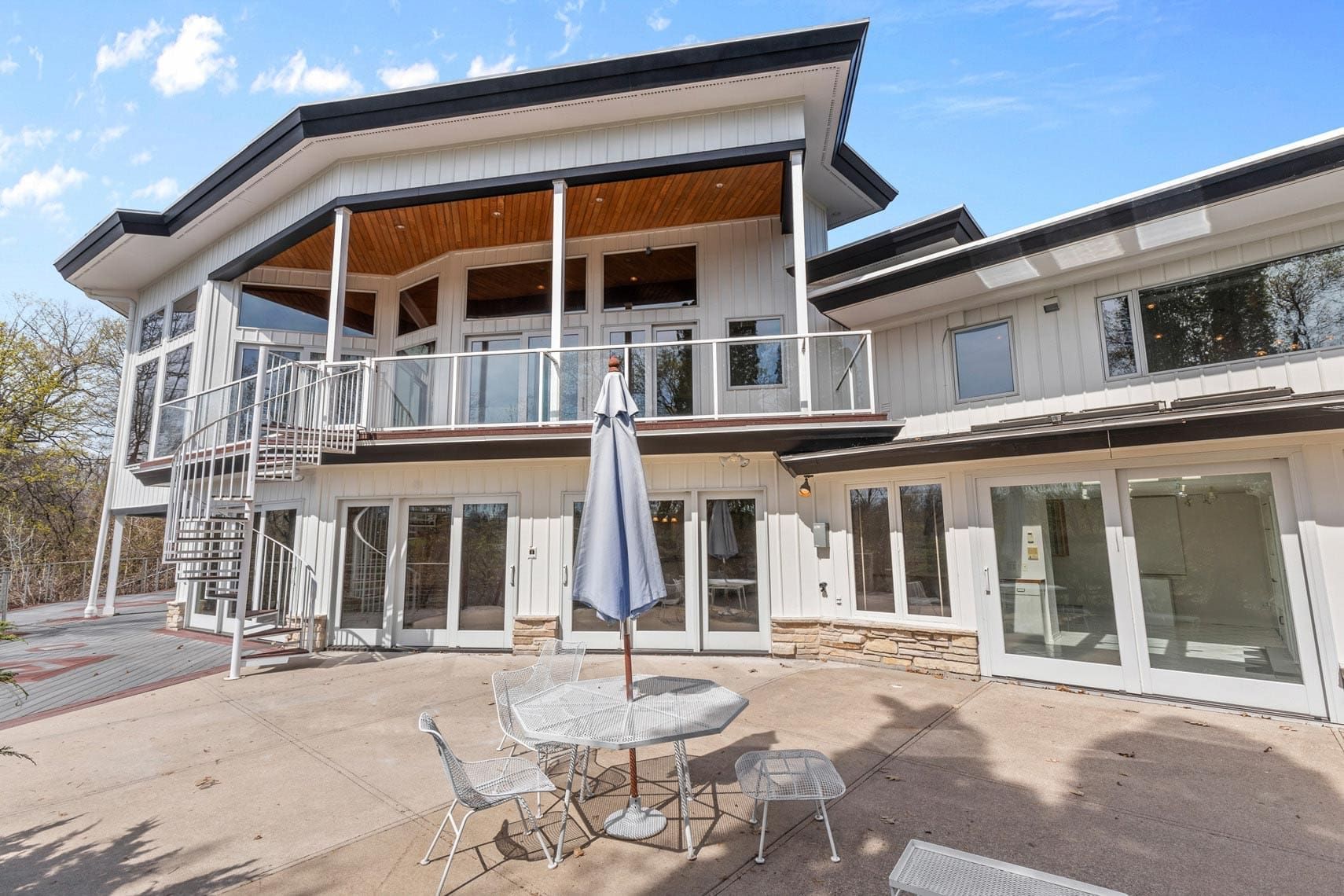 Spacious modern residential property featuring large windows, an upper balcony, and a stylish spiral staircase. Commercial real estate image showcasing an outdoor patio area with a table, chairs, and a closed umbrella beneath a clear blue sky. Ideal for vacation rental promotions.