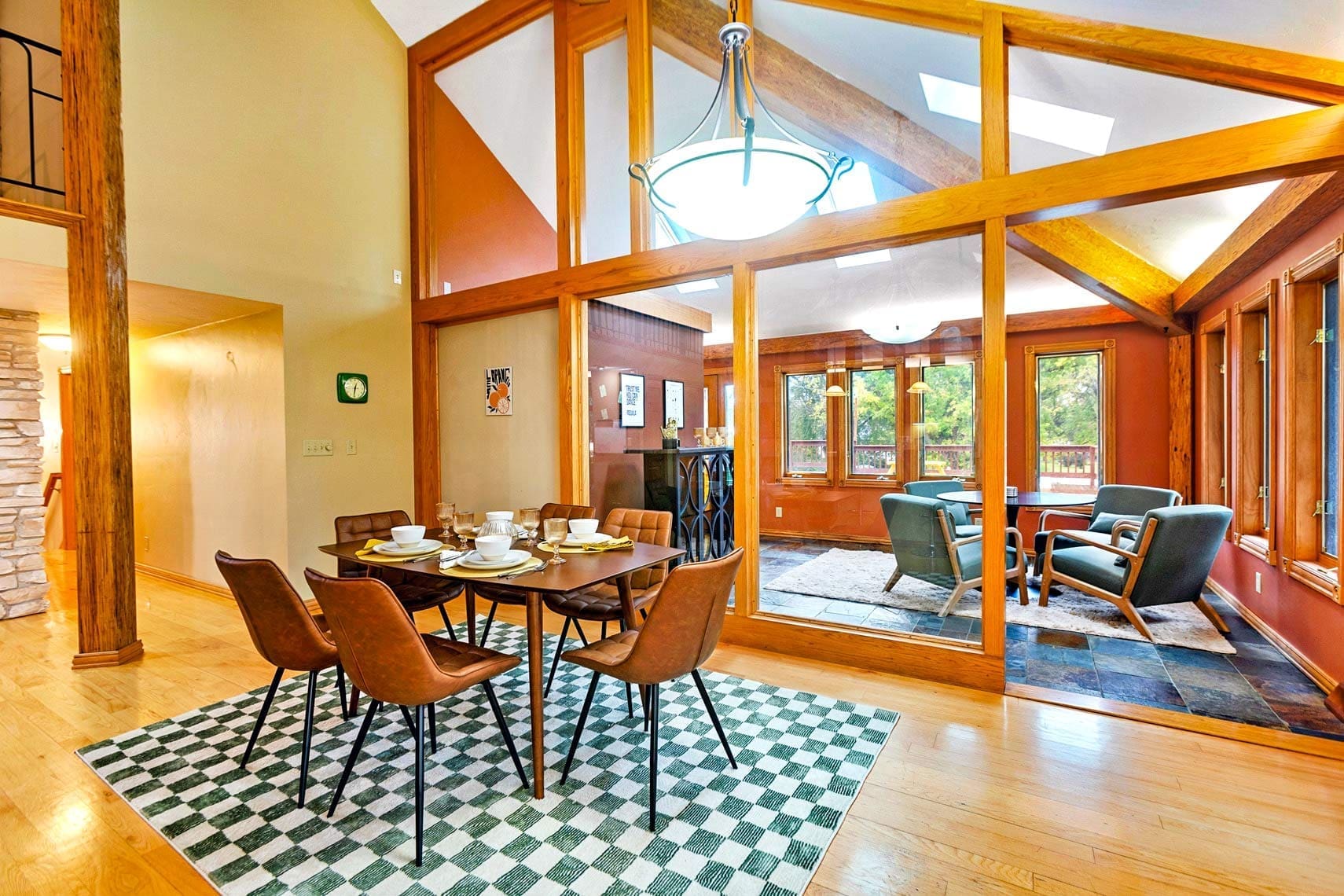 Residential dining room with a wooden table and chairs on a checkered rug, next to living room with armchairs and large windows; featuring wood beams and vaulted ceiling, ideal for real estate photography.