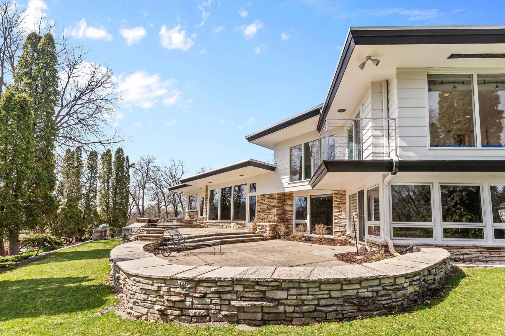 Contemporary residential property with expansive windows and a stone patio, nestled in lush landscaping under a partly cloudy sky.