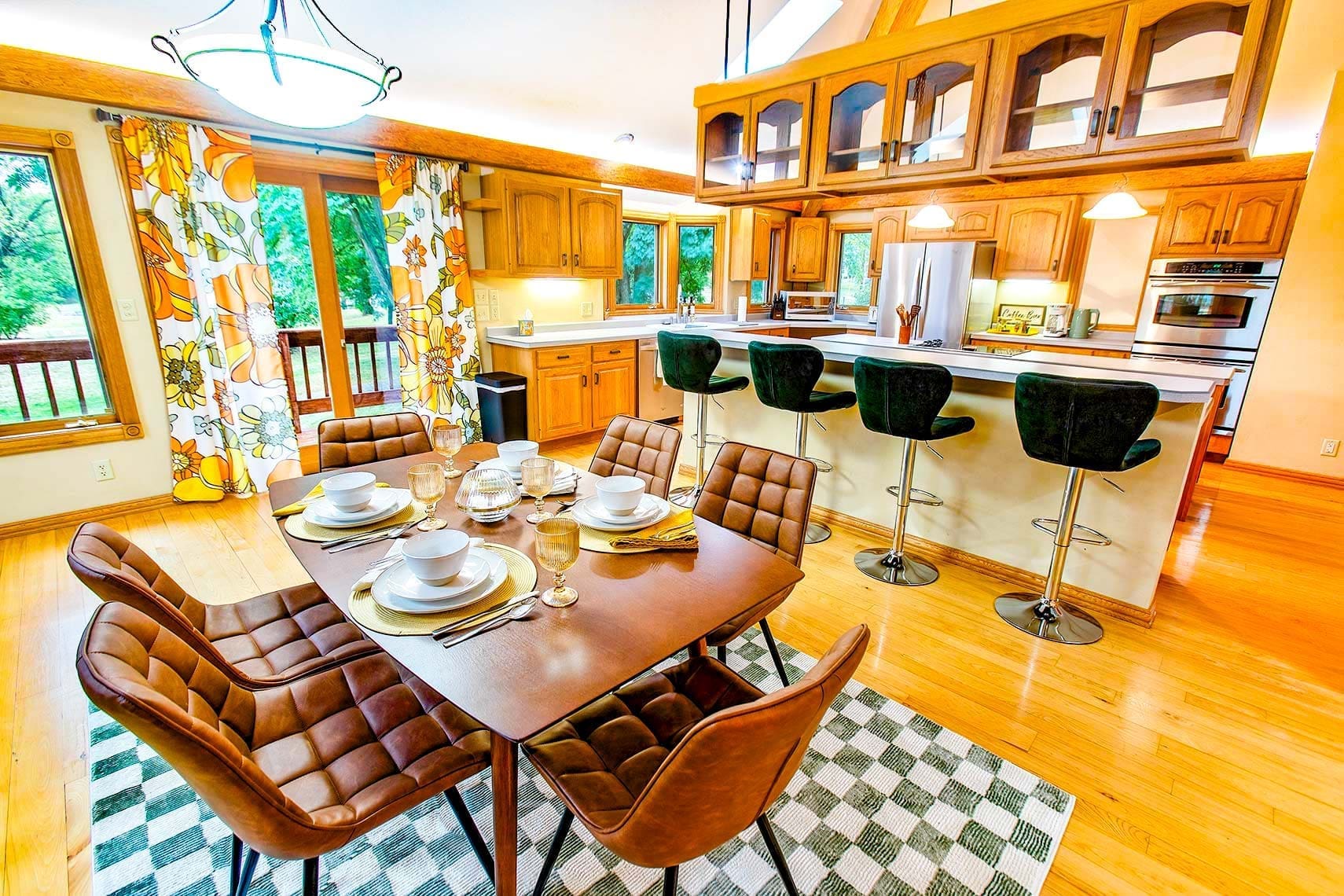 Modern residential kitchen and dining area with wooden cabinetry, dining table set for six, green velvet bar stools, floral curtains, and scenic greenery view through windows.