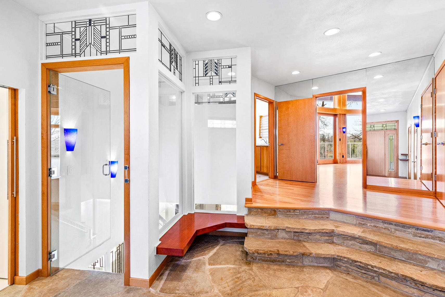 Modern residential interior showcasing wooden and stone elements, with a glass door, geometric windows, and steps leading to an open hallway. Bright lighting enhances the minimalist design in this real estate property photo.