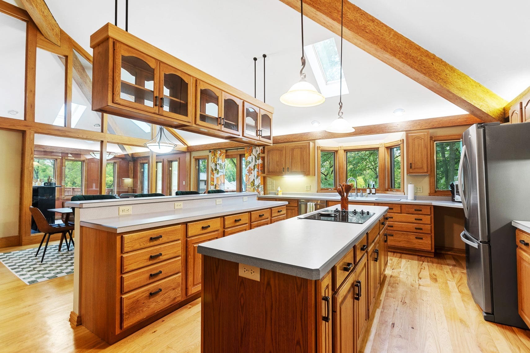 Spacious kitchen in residential property featuring wooden cabinetry, center island with stove, stainless steel refrigerator, and pendant lights; ideal for real estate photography. Large windows and skylight allow natural light to flood the space. Dining area in background highlights open-concept design.
